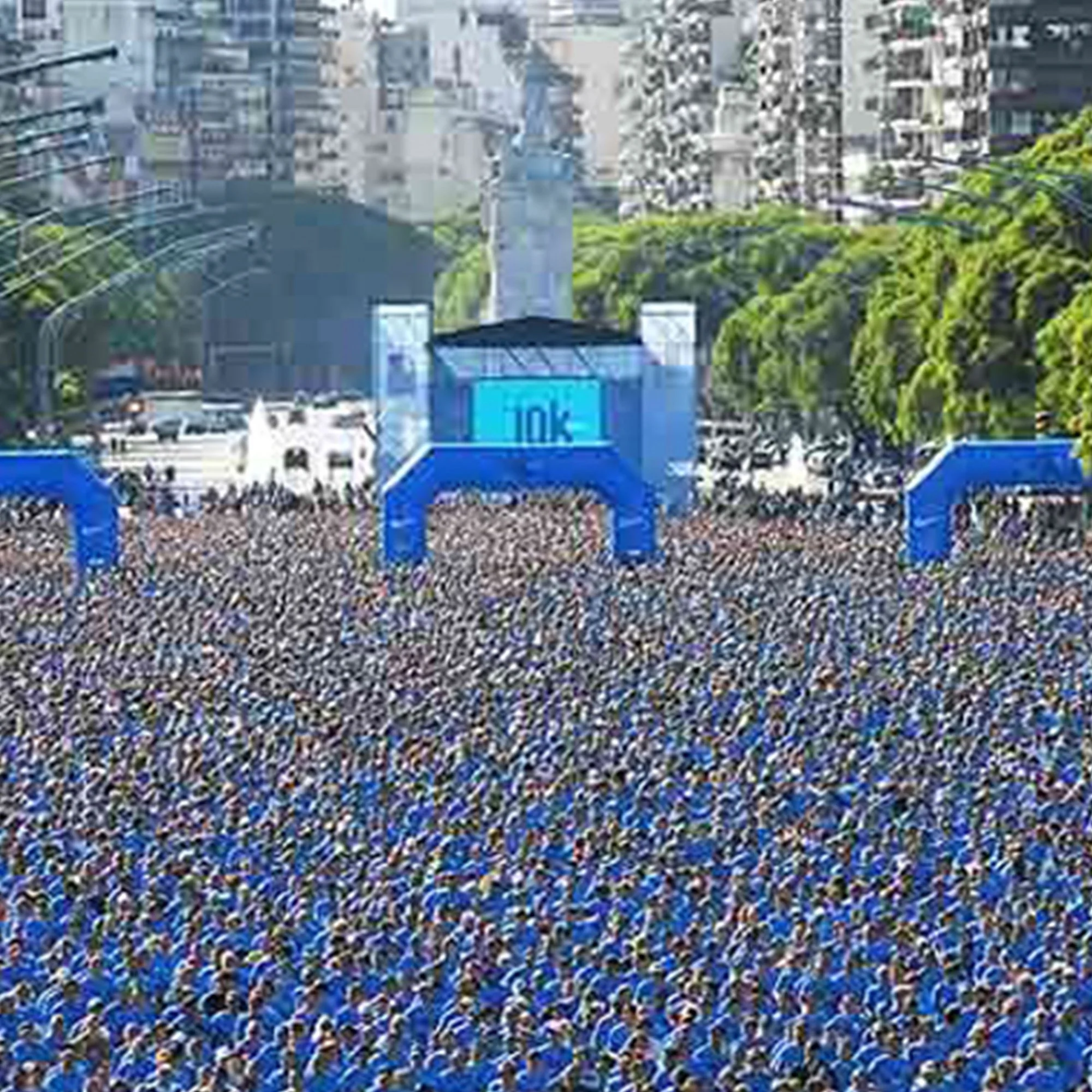 A large crowd of people walking along a city street with high-rise buildings and trees, with a blue structure labeled 'Link' in the foreground.