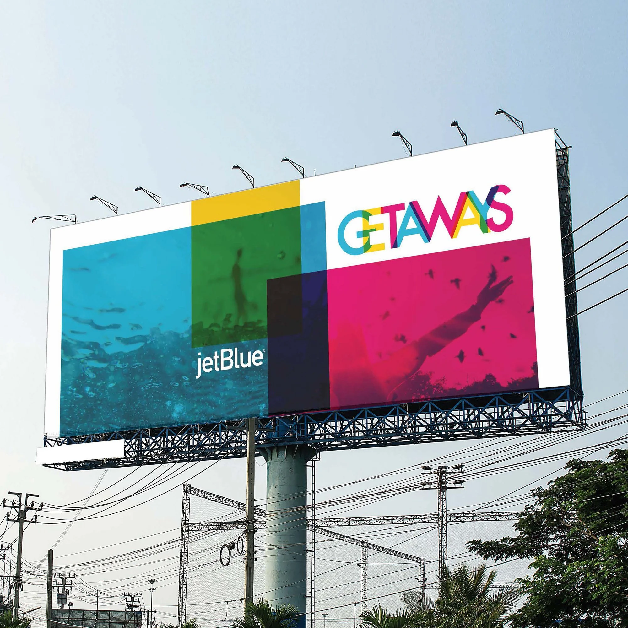 A large colorful billboard with the words 'GETAWAYS' and the JetBlue logo underneath, mounted on a metal structure above power lines and trees in an outdoor setting.