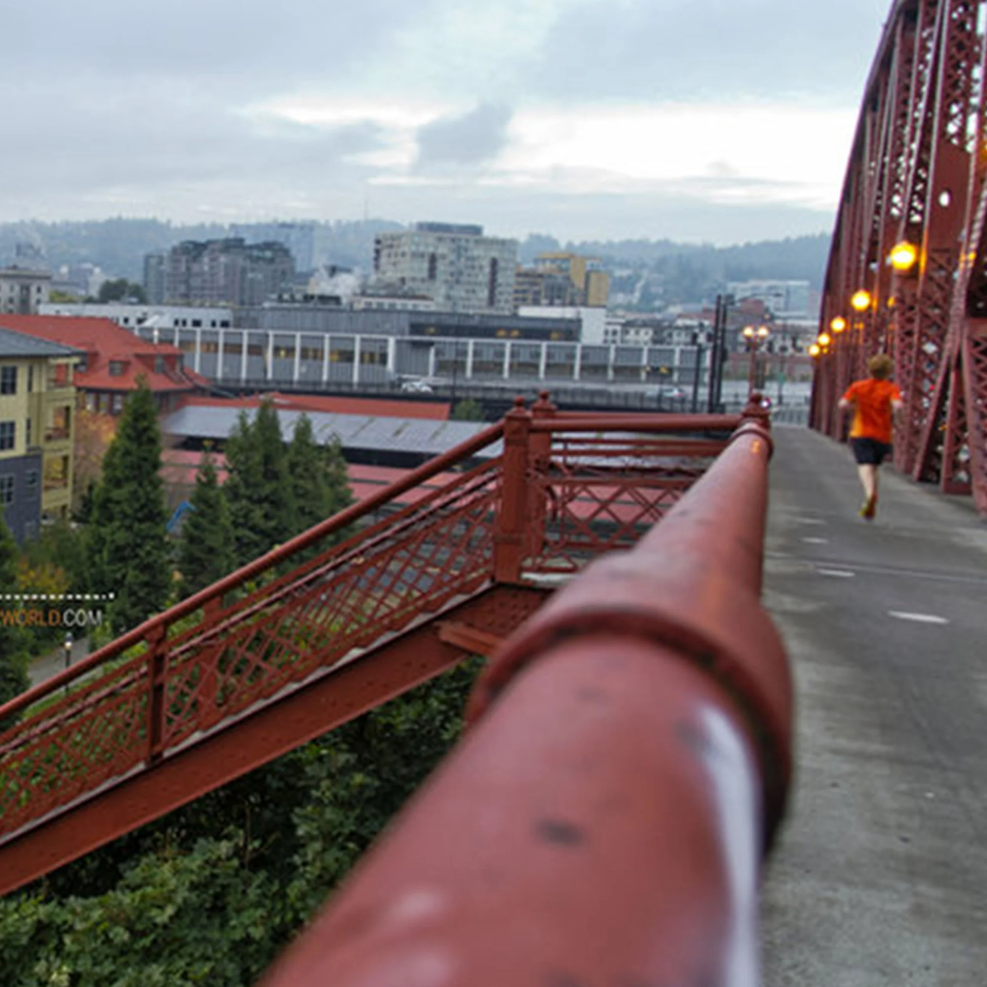 View of a city skyline from a red bridge with string lights, overcast sky, and a person walking on the bridge.