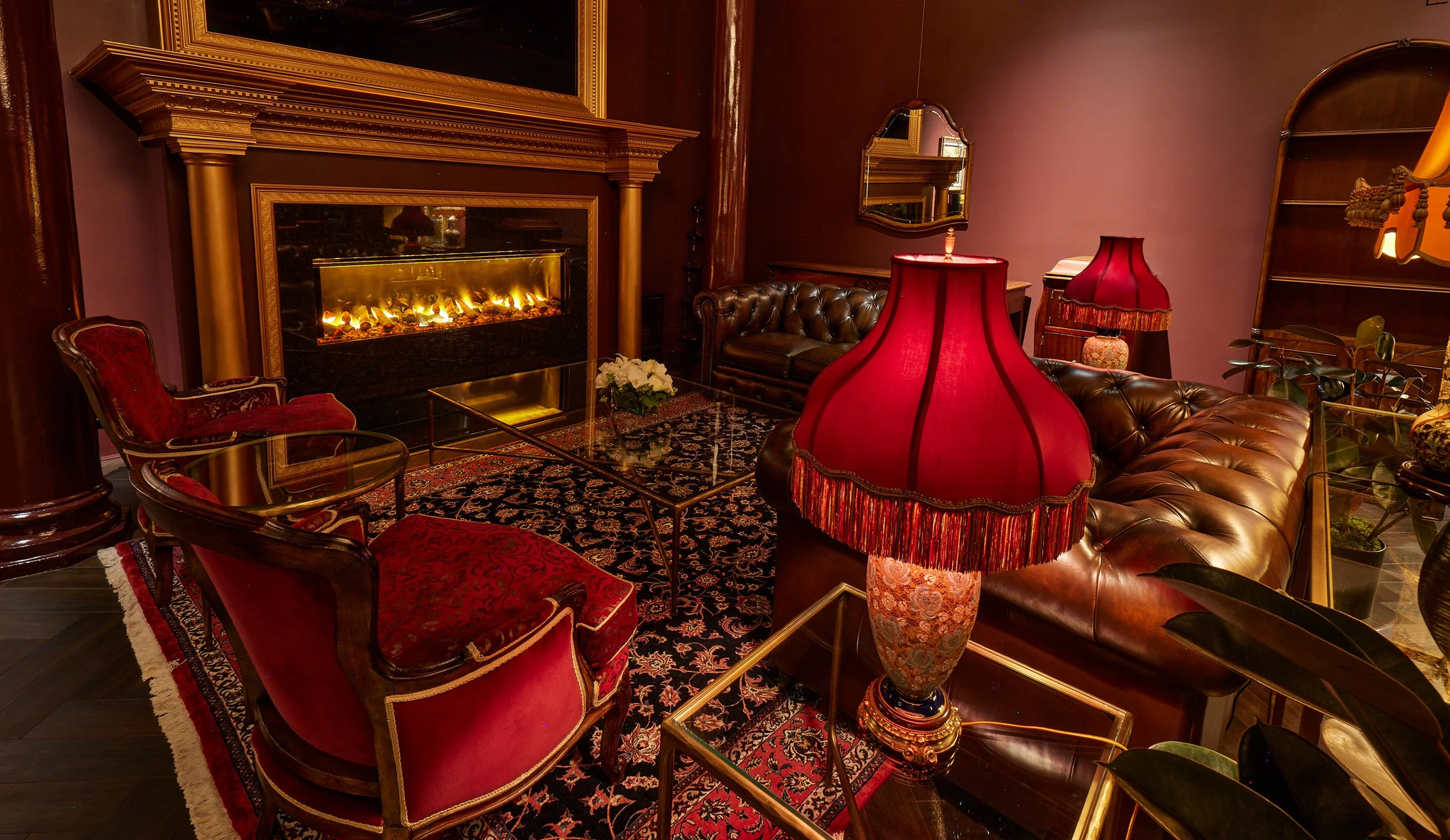 A cozy living room with a fireplace, brown leather sofas, red velvet chairs, ornate lamps, a mirror, and various decorative items including a plant and a patterned rug.