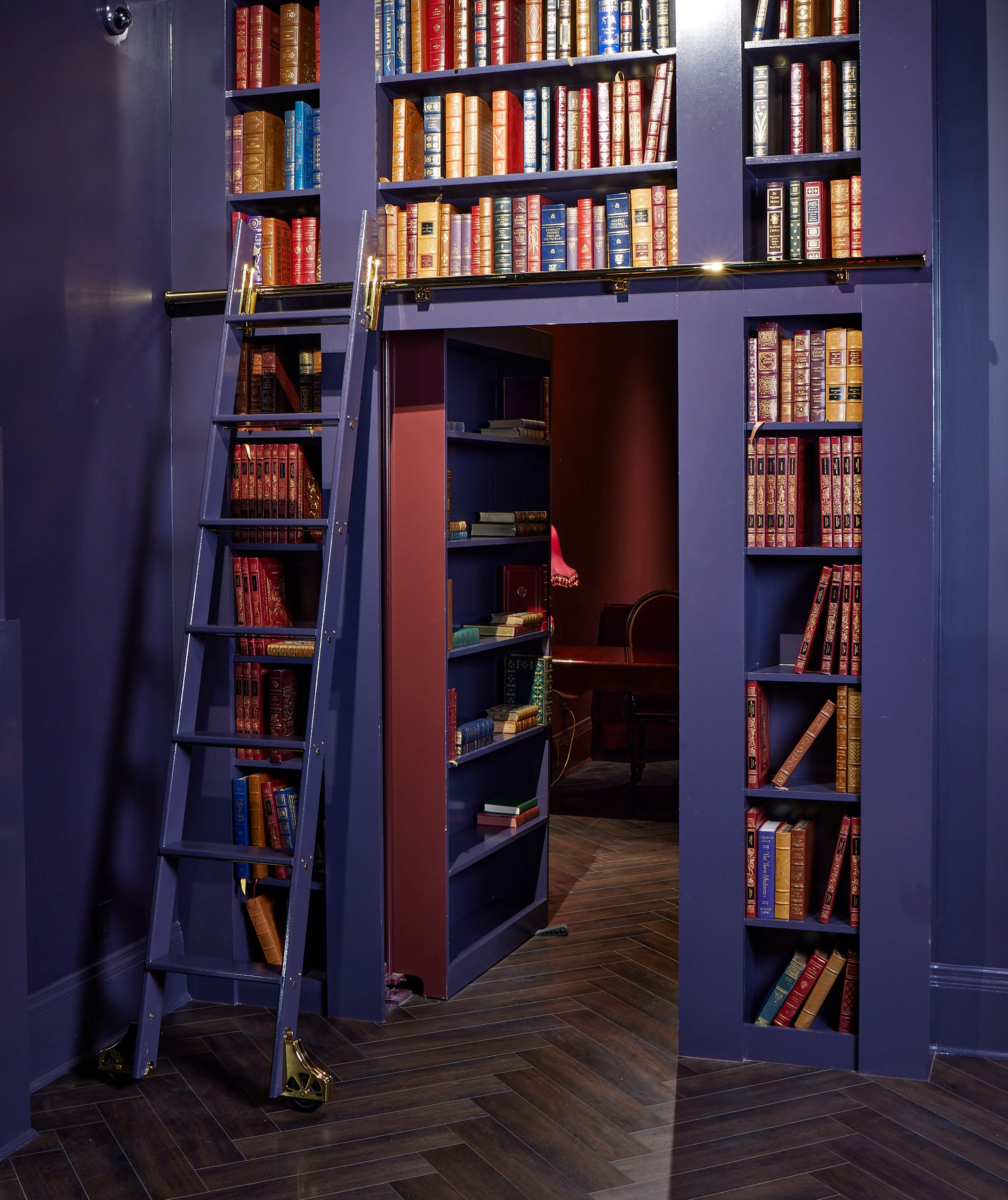 A library room with dark blue bookshelves filled with leather-bound books, a ladder with gold high heels at the bottom, and wooden floor.