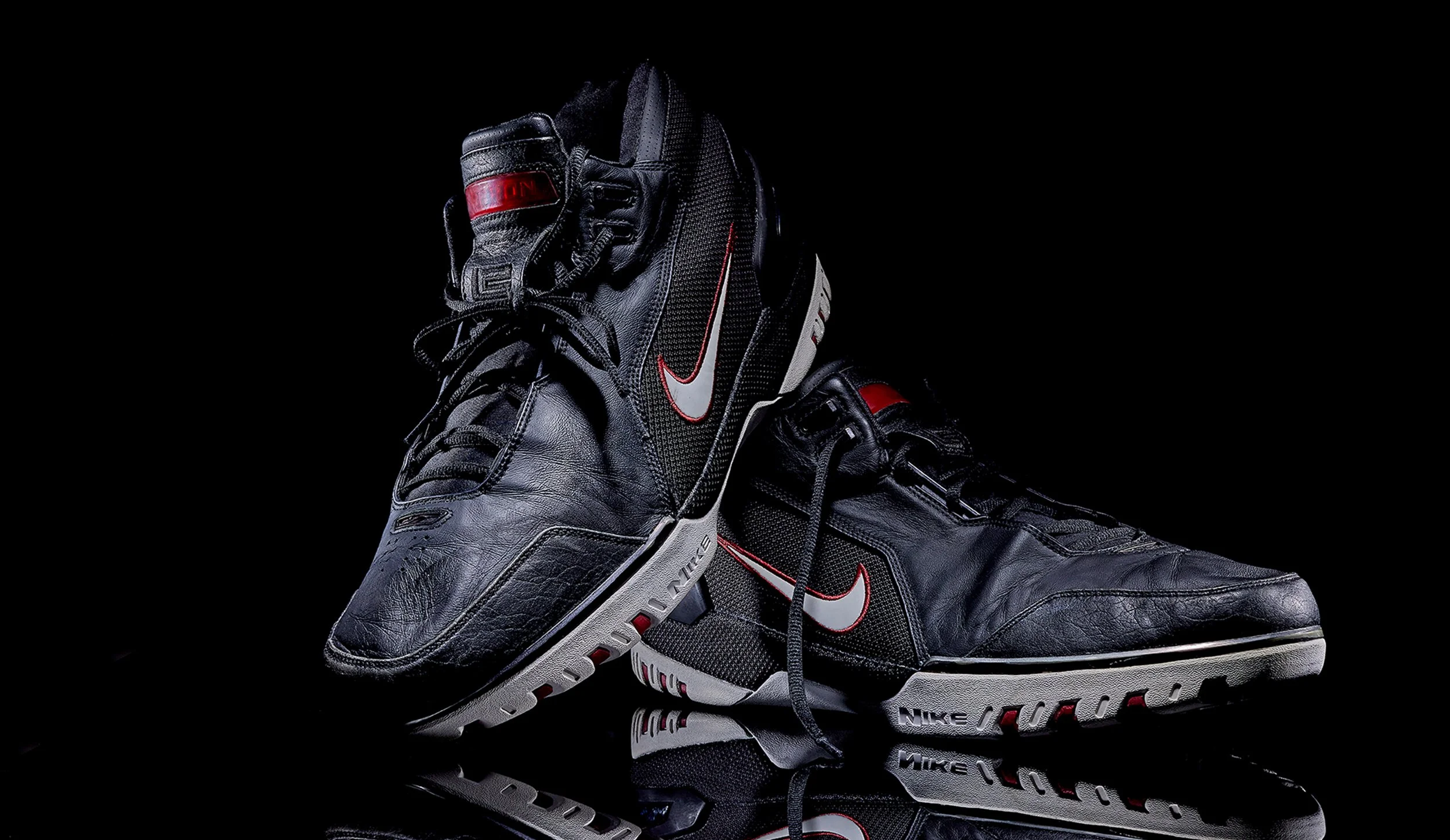 A pair of black Nike Air Jordan sneakers with red and white accents, positioned on a reflective surface against a black background.