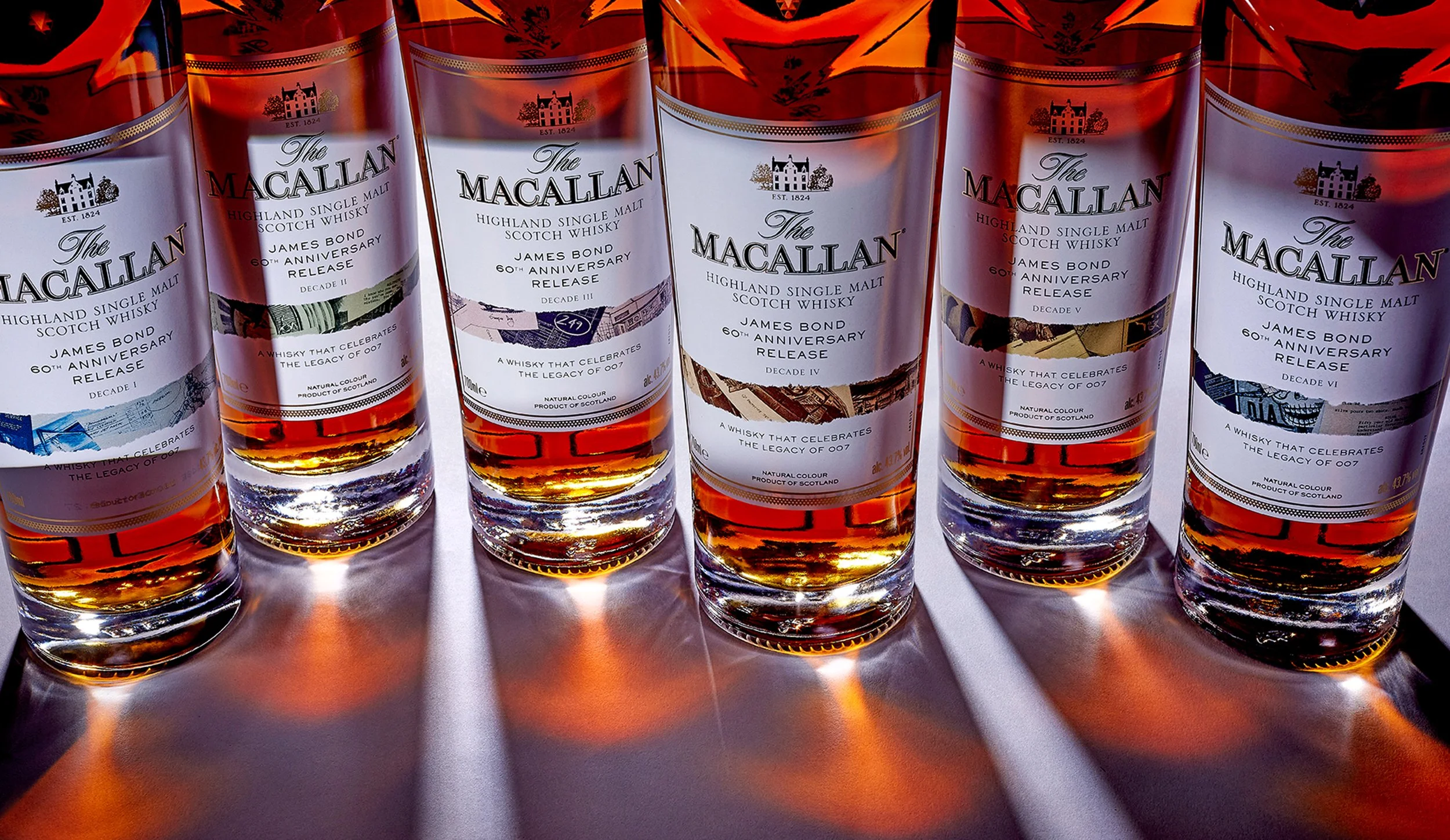 Showcase of multiple bottles of The Macallan Highland Single Malt Scotch Whisky in a row, each with a white label celebrating a James Bond 60th anniversary release, varying in Decades from I to VI.