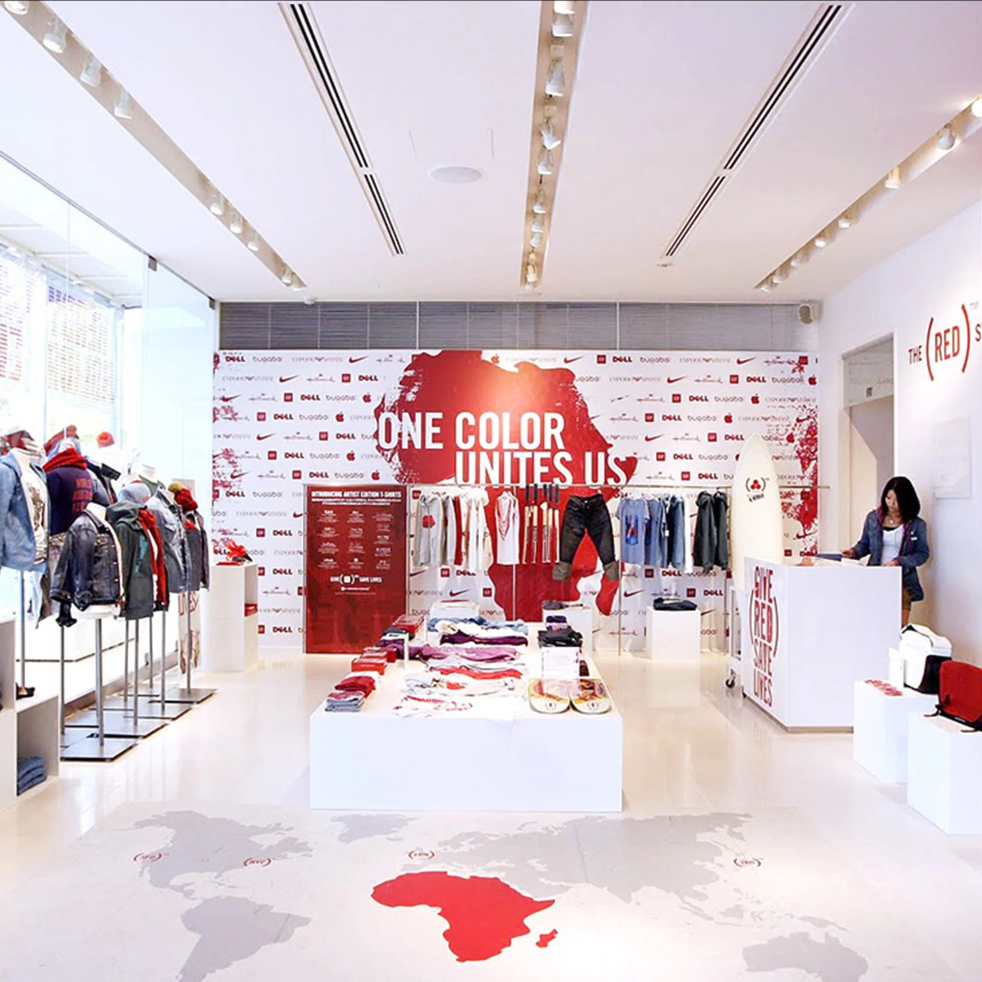 Retail store interior with white walls, display shelves of shoes and clothing, and a front area featuring a large red, white, and black banner with the words "ONE COLOR UNITES US" and an outline of the country of New Zealand.