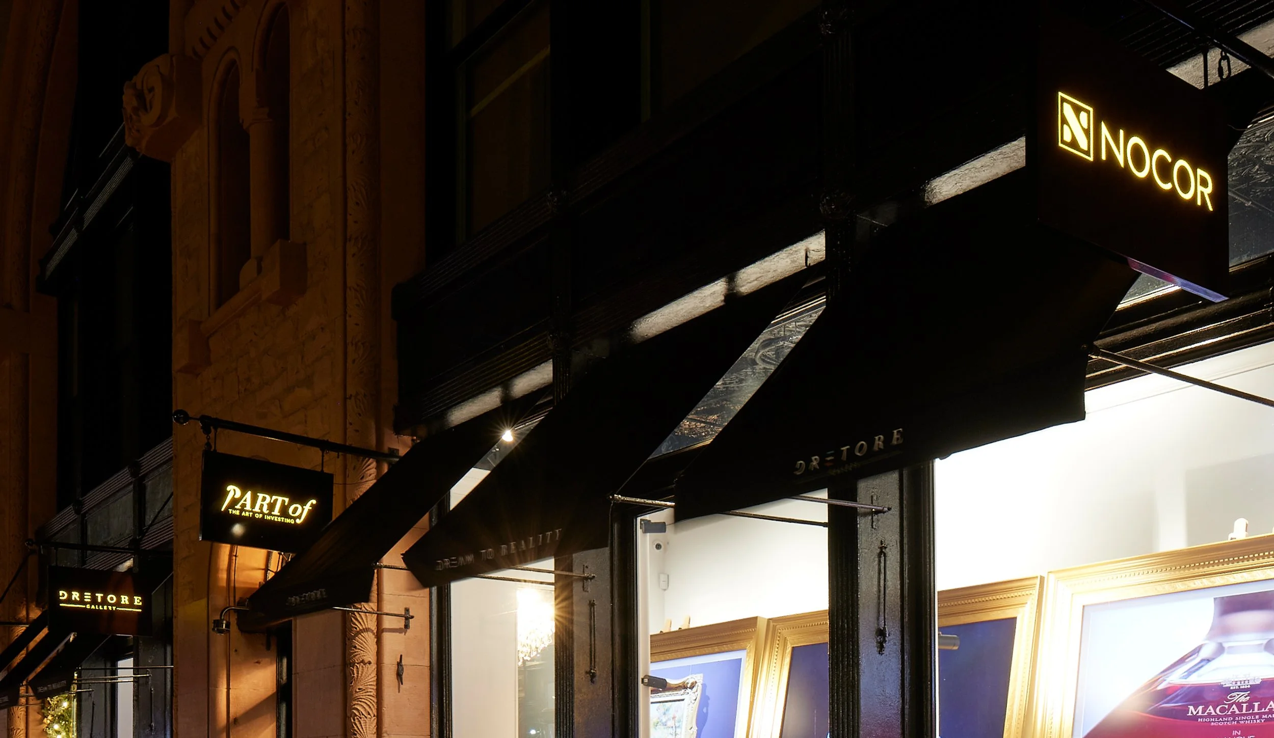 Night view of storefront signs including 'NO COR' and 'PART OF THE ART OF INVESTING' on a building with ornate architectural details, illuminated by warm lighting.