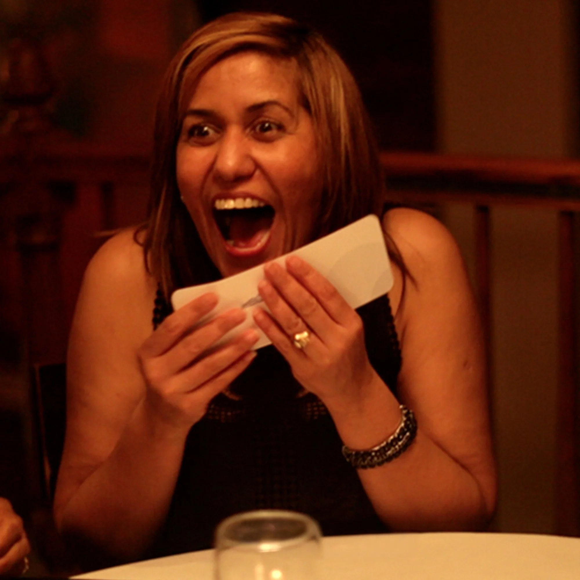 Close-up of a woman with brown hair smiling and showing her teeth, holding a white cup, sitting at a table in a dimly lit setting.