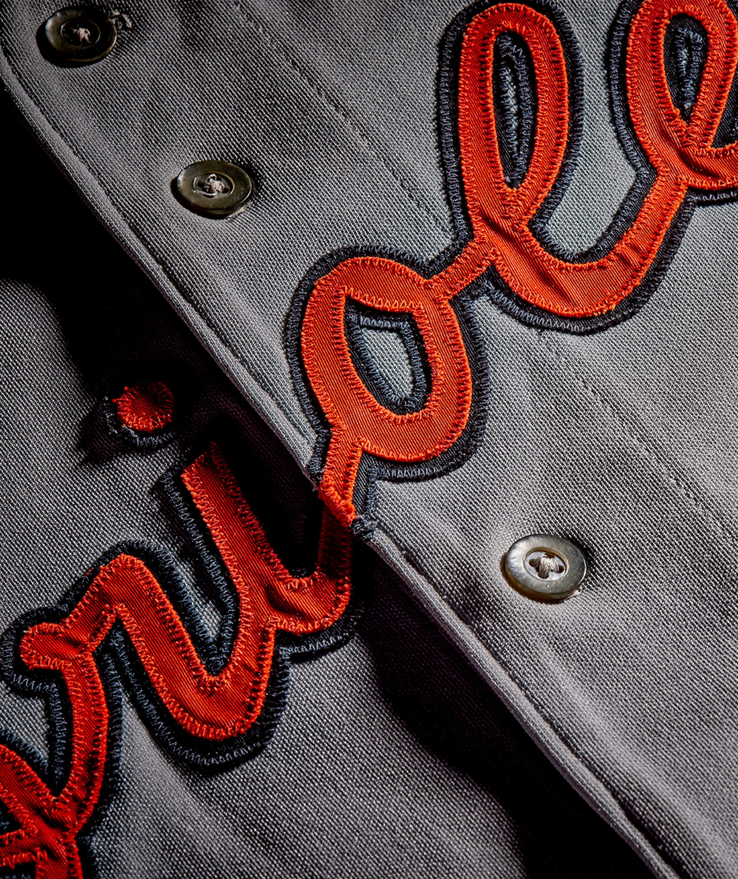 Close-up of a gray fabric jacket with black buttons and embroidered orange and black text and design.