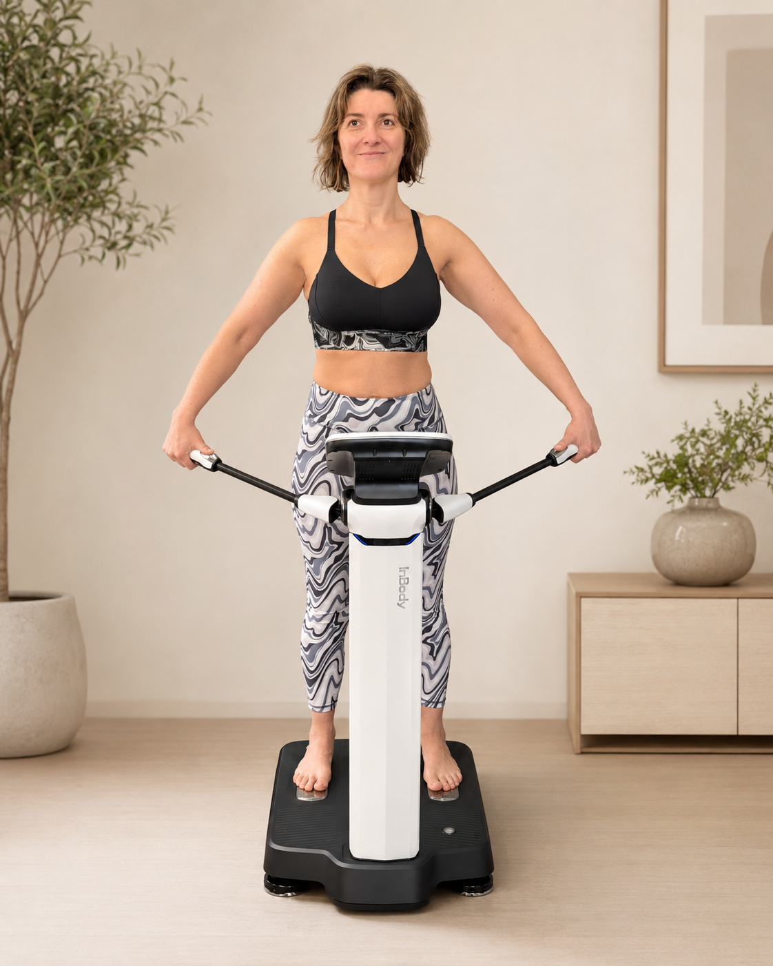 Woman standing on a digital wellness platform, holding handlebars, wearing workout clothing, in a room with potted plants and a framed picture on the wall.