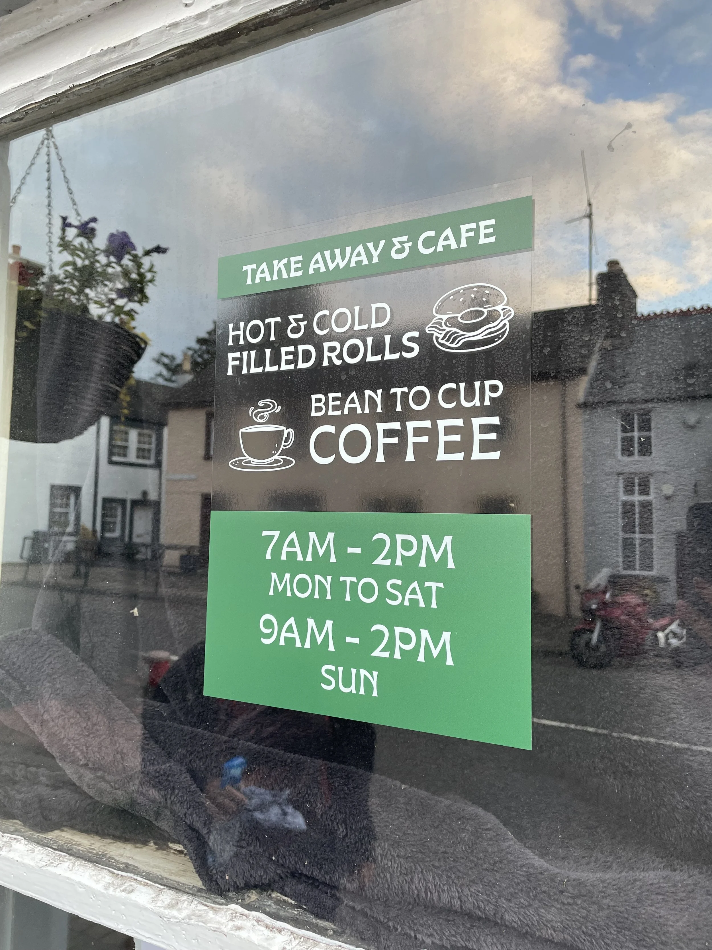 Sign on a café window advertising take away and cafe options including hot and cold filled rolls, bean to cup coffee, with operating hours from 7 am to 2 pm Monday to Saturday and 9 am to 2 pm on Sunday.