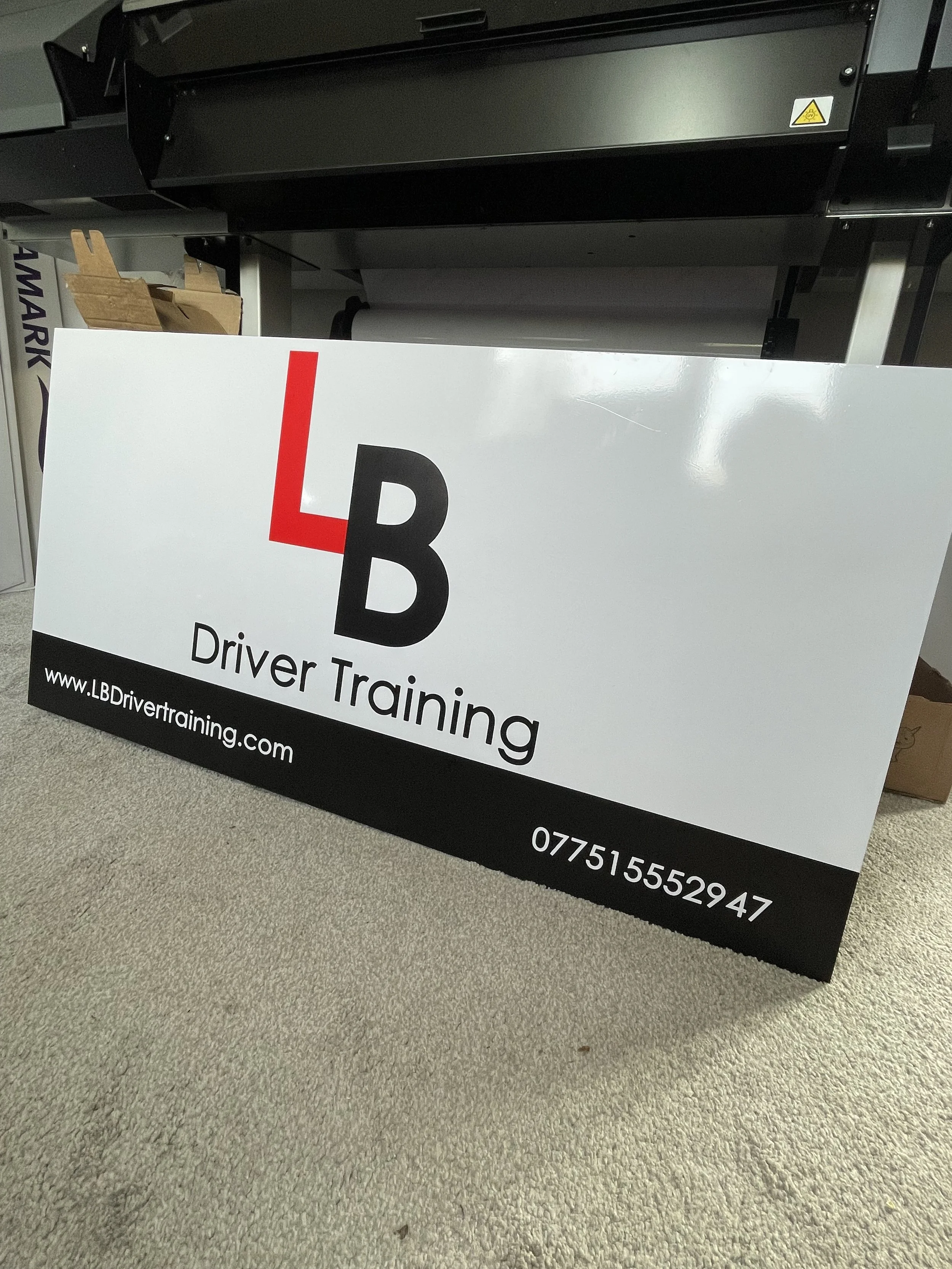 A large white sign with black and red text advertising LB Driver Training, including a website and phone number, placed on a carpeted floor in front of a black machine or printer.