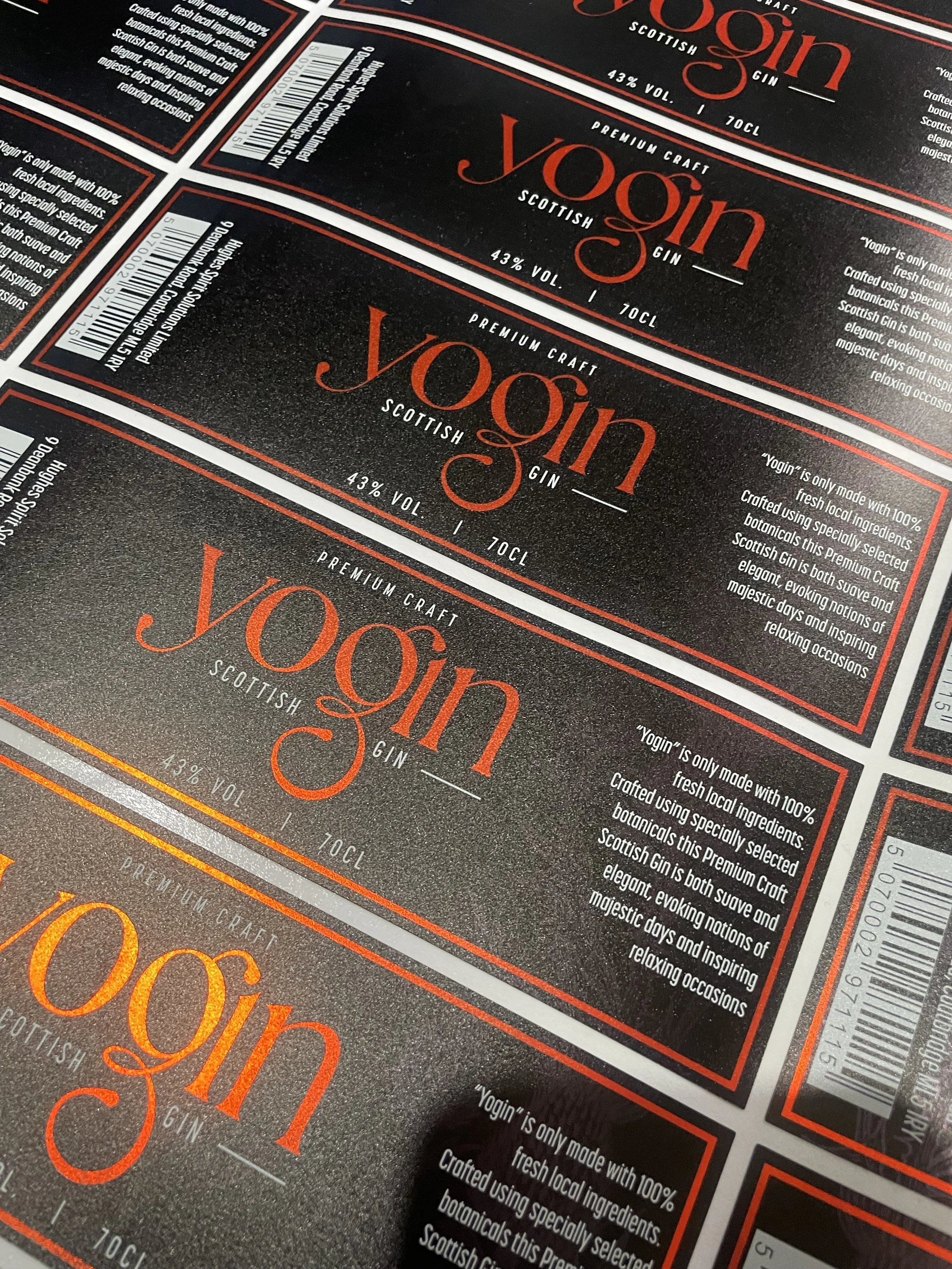 Close-up of multiple labels for 'Yogin Scottish Gin,' featuring the brand logo in large orange text, alcohol details, and a paragraph describing the gin's qualities and ingredients.