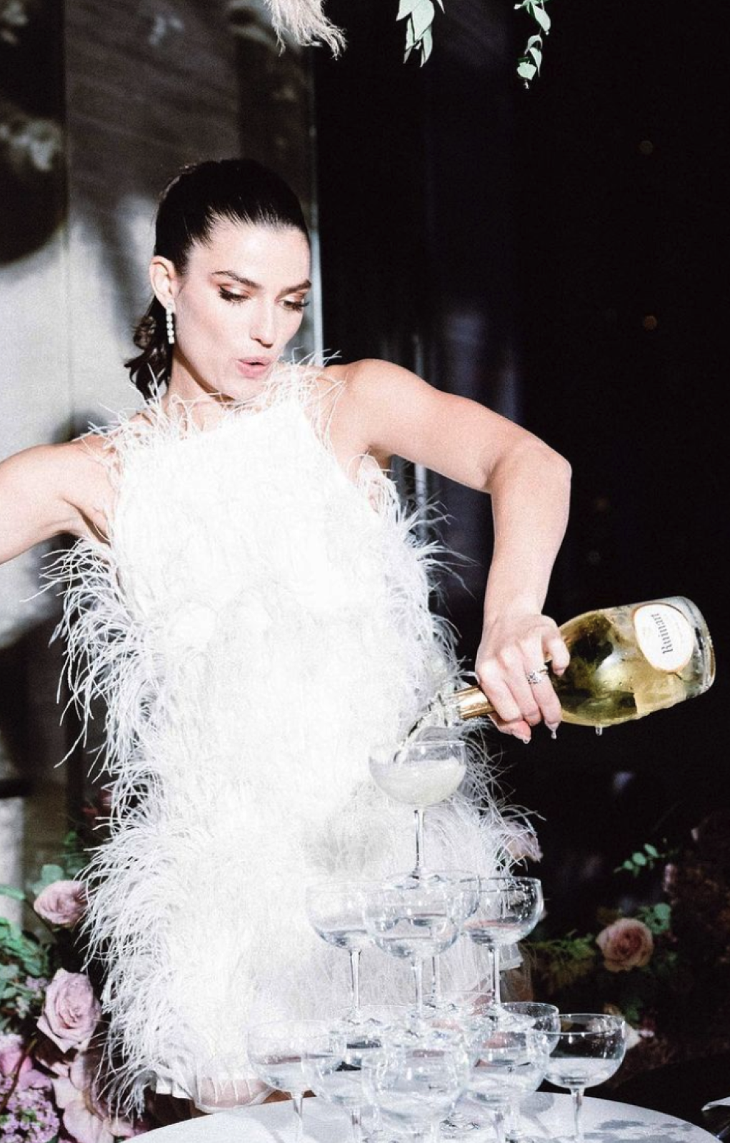 A woman in a white feathered dress is pouring champagne into a tower of glasses.