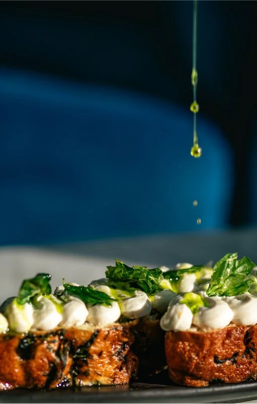 Two baked appetizers topped with white sauce and fresh green herbs, with droplets of oil falling from above.