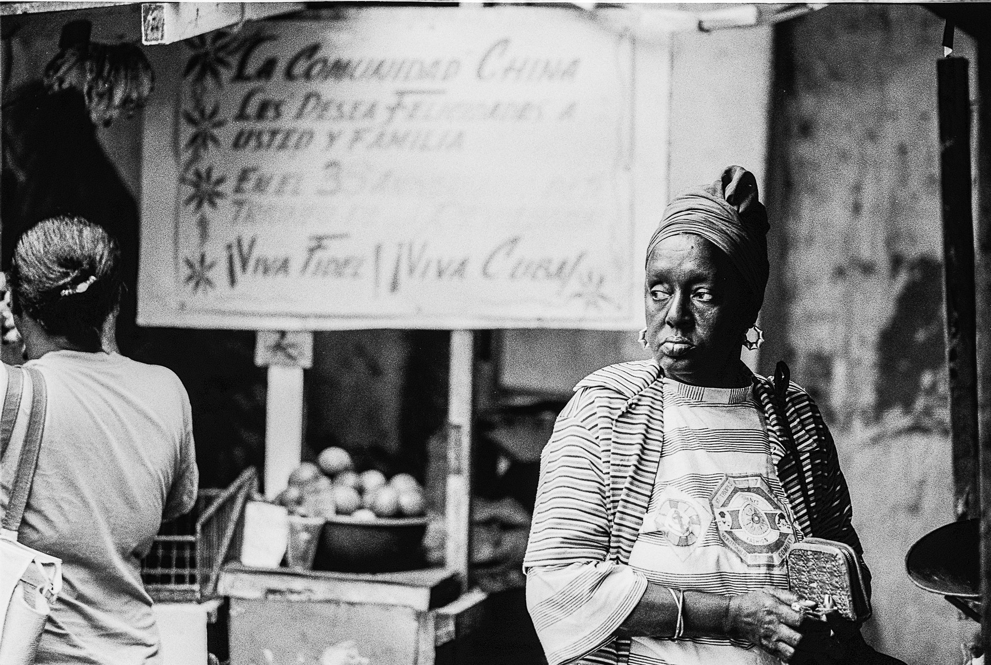 A woman wearing a headscarf and patterned shirt stands in front of a blurred sign at an outdoor market, holding a small purse. Other people and produce are visible in the background.