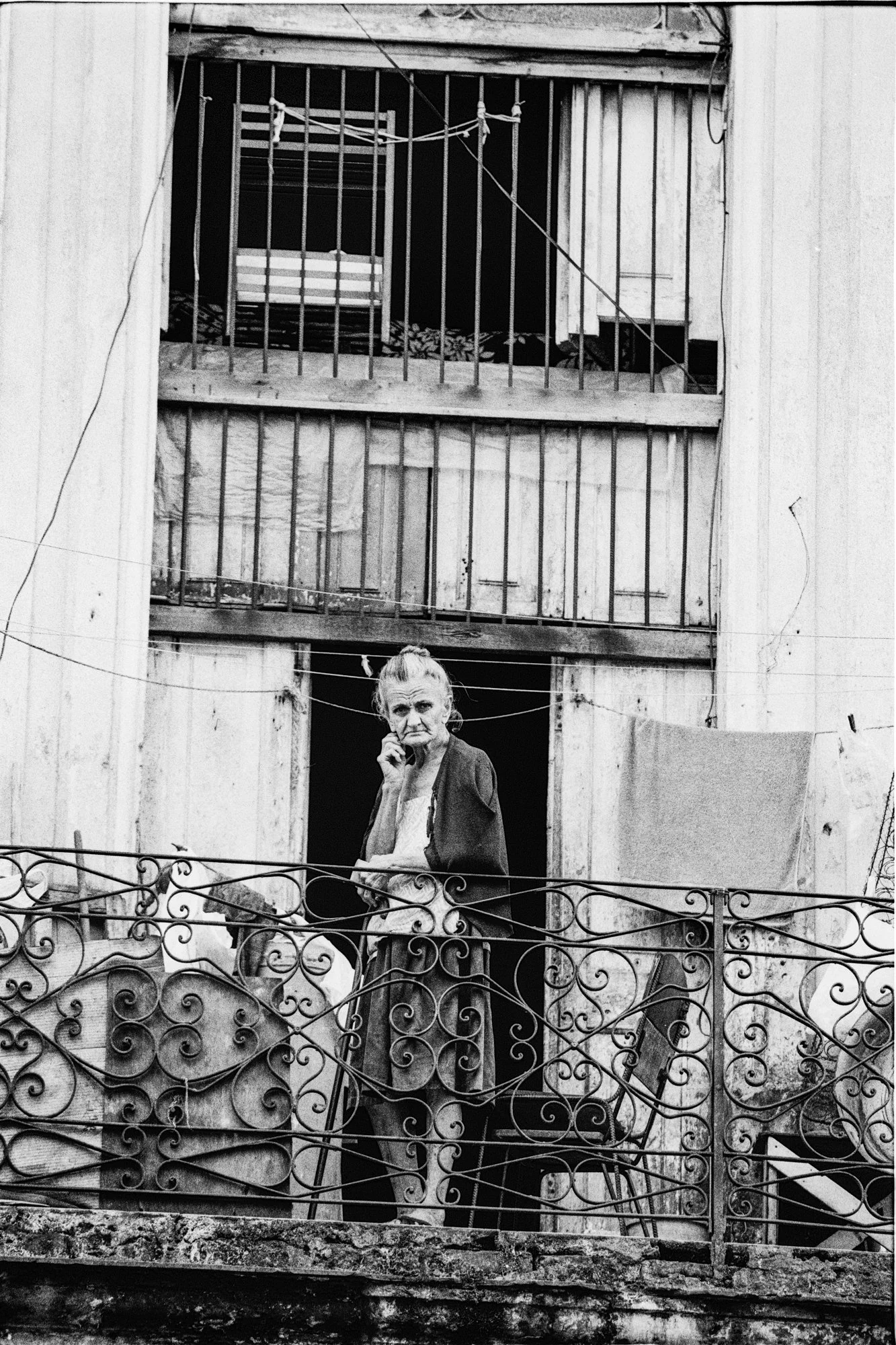 An elderly woman standing on a balcony with ornate metal railings, talking on a phone, with a worn building behind her.