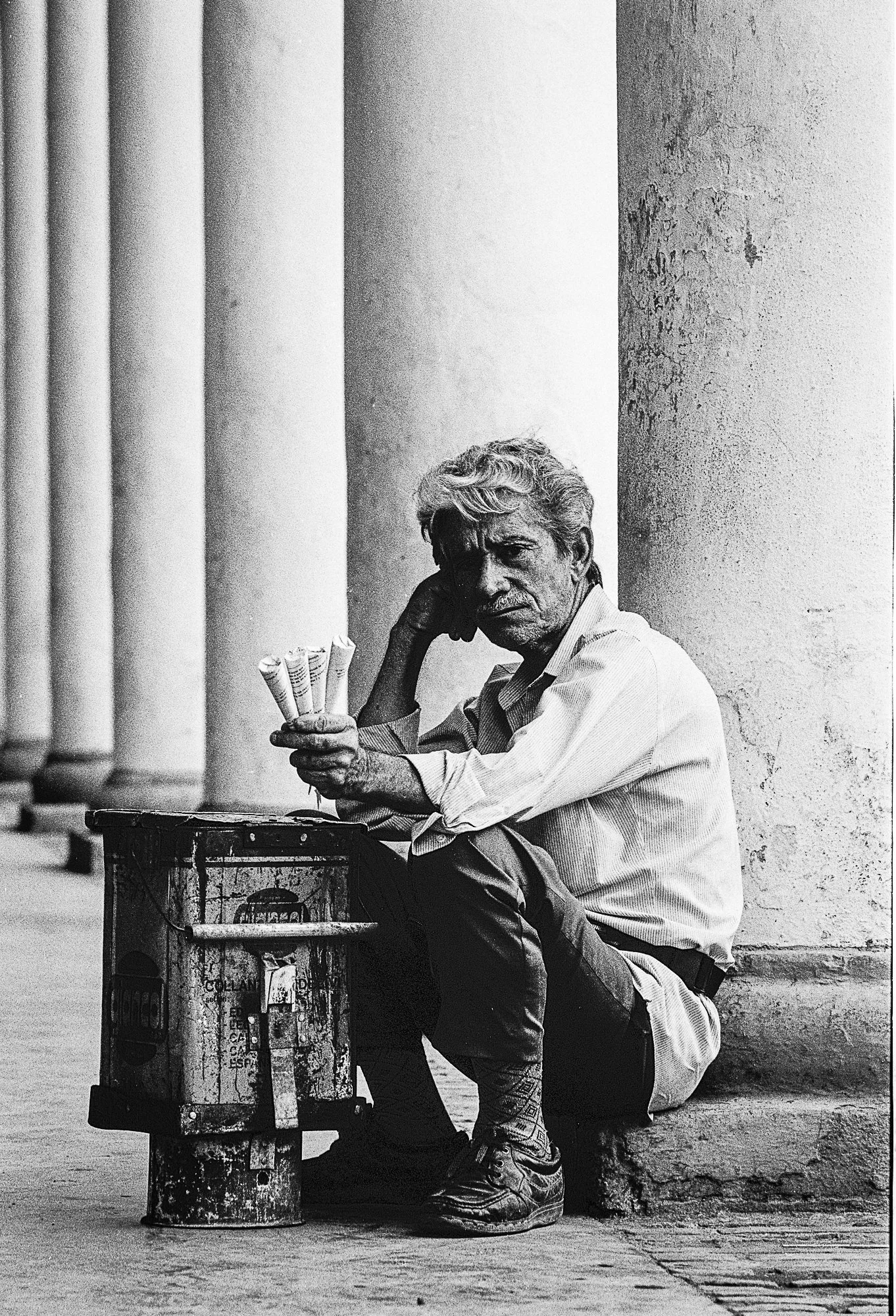 Black and white photograph of a man sitting on the sidewalk, leaning against a large column. He is holding a bundle of newspapers or papers, with a small, makeshift stove or heater in front of him. The man appears to have gray, curly hair and is wear