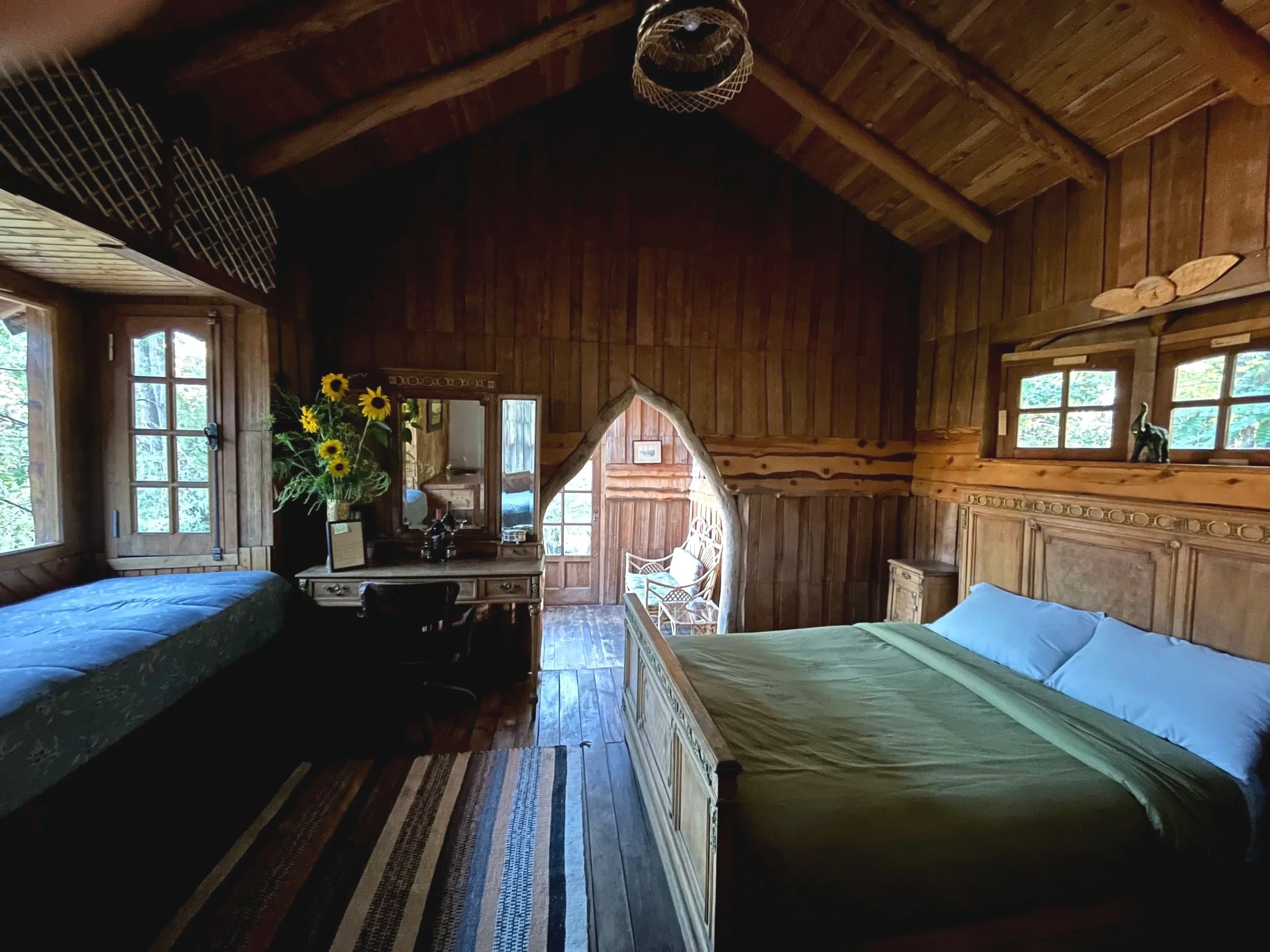 A rustic bedroom with wooden walls, ceiling, and floors, featuring a bed with green bedding, a window, a dresser with a mirror, a sunflower bouquet, and a small sitting area outside through a doorway.