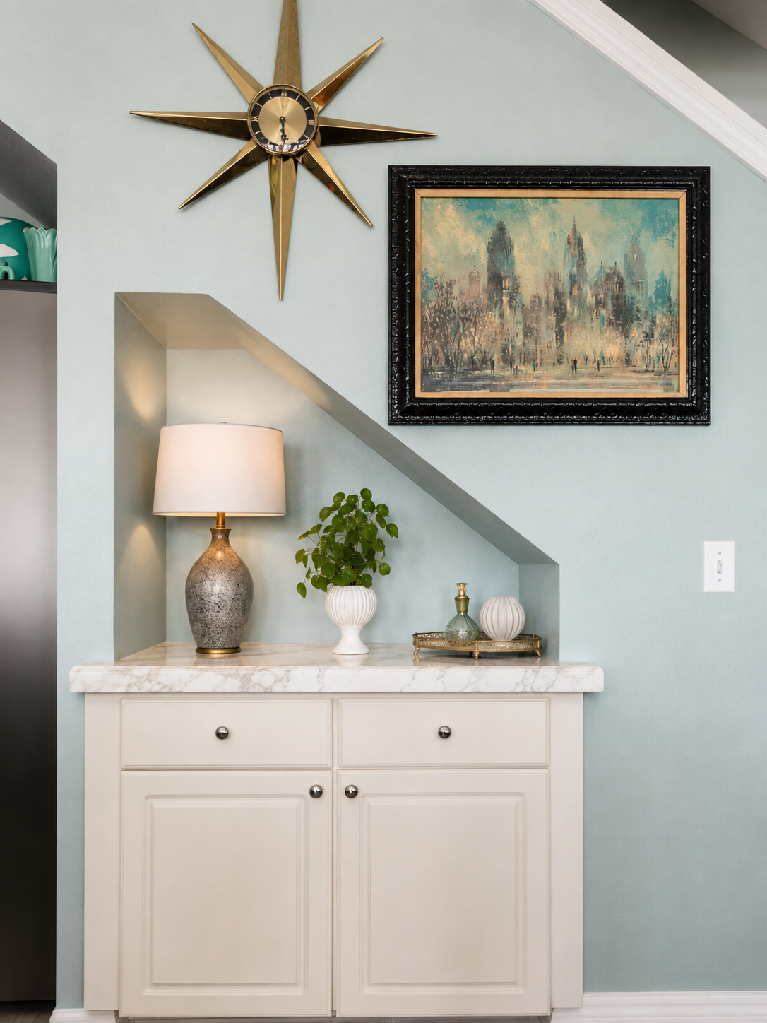 Vancouver Condo in the Portland Metro Area designed by Linda McDermott of McDermott Interiors, cabinet with a marble top, decorative table lamp, potted plant, ceramic jars, modern wall clock, and framed artwork against a soft blue wall.