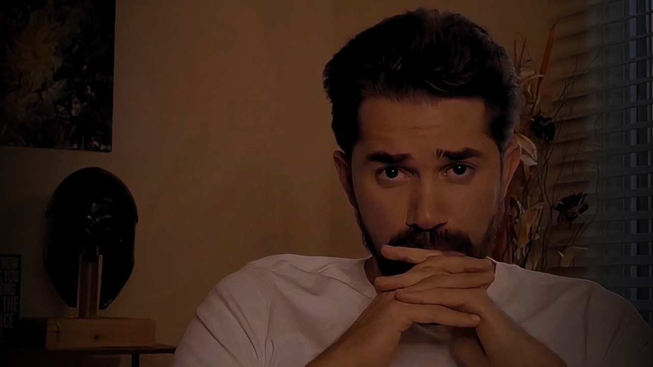 A man with dark hair and a beard sitting indoors, resting his chin on his hands, looking towards the camera with a serious expression. Background includes a window with blinds and some plants.