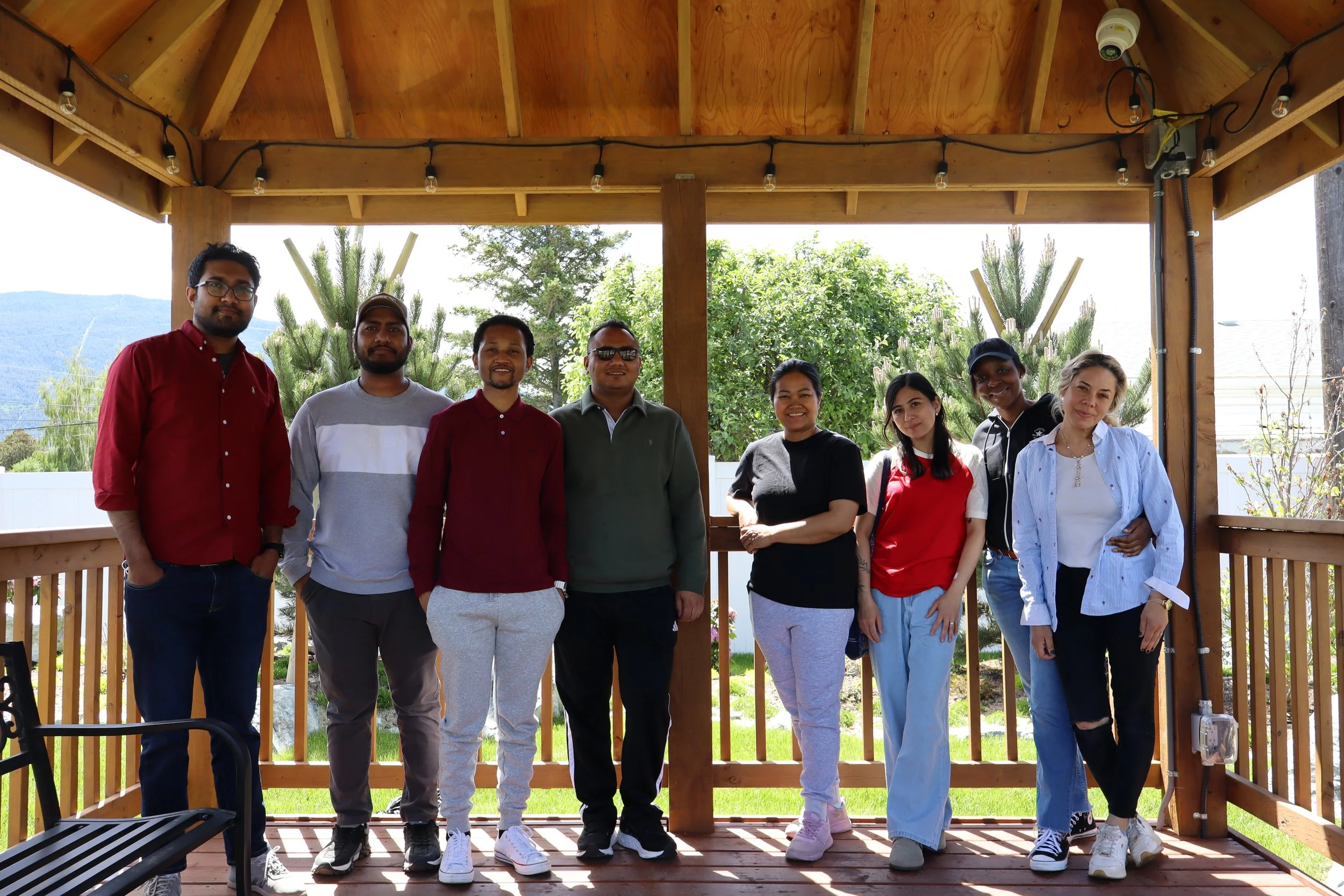 Group of eight diverse young adults standing on a wooden deck with a lush green backyard background, under a wooden roof with string lights and security cameras.