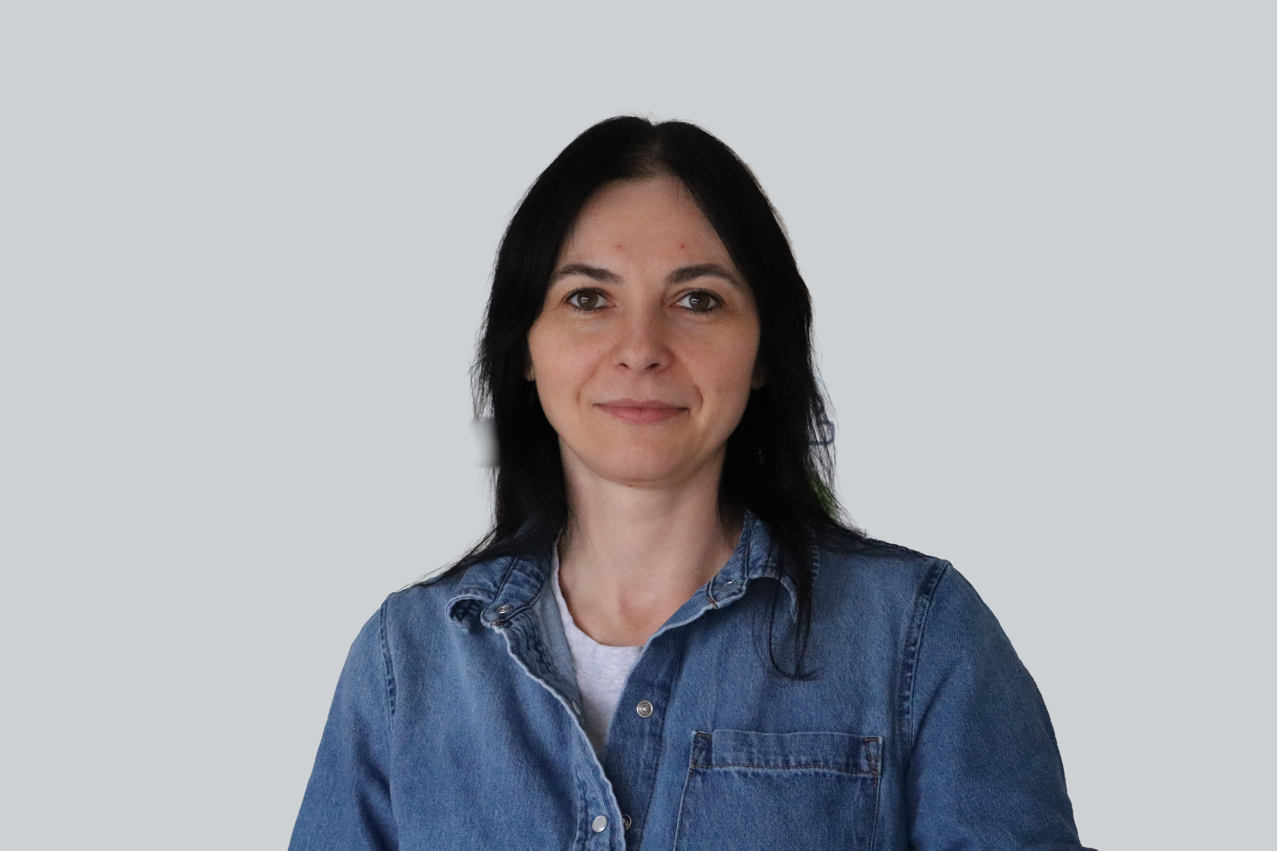 A woman with straight black hair, wearing a blue denim shirt over a white t-shirt, standing against a plain light grey background.
