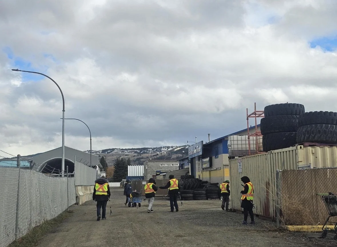 Group of people wearing reflective vests walking in an industrial yard with stacked tires and buildings in the background under cloudy sky.