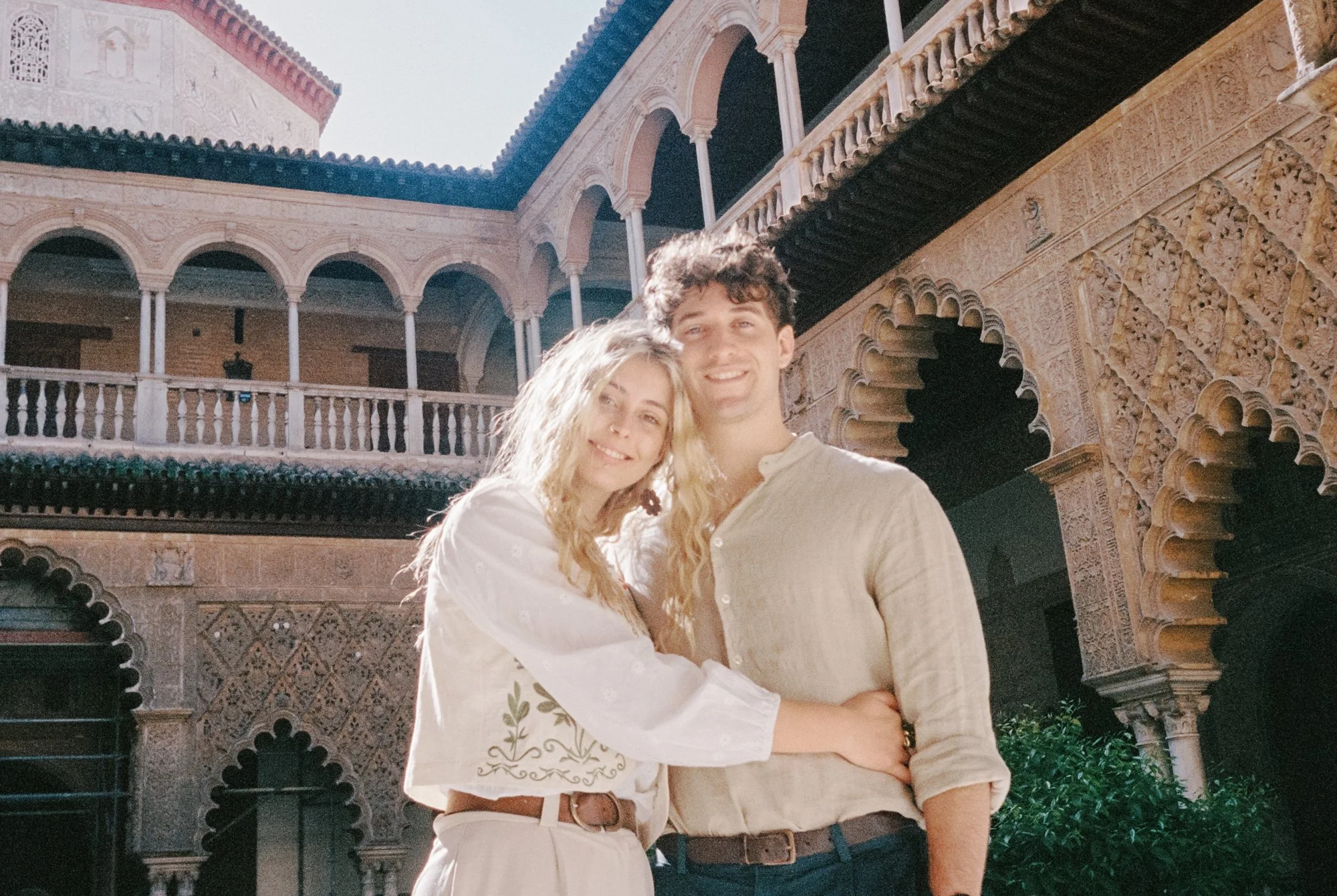 A smiling young couple who are a wedding filmmaking team, stands close together outside a historic building with intricate architectural details, arches, and balcony railings, illuminated by bright sunlight.