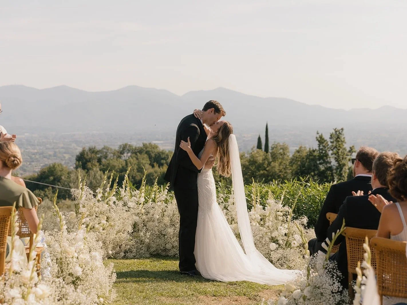A bride and groom share a kiss during their outdoor wedding in a scenic rural setting with mountains in the background, surrounded by guests seated on wooden chairs and decorated with white flowers.