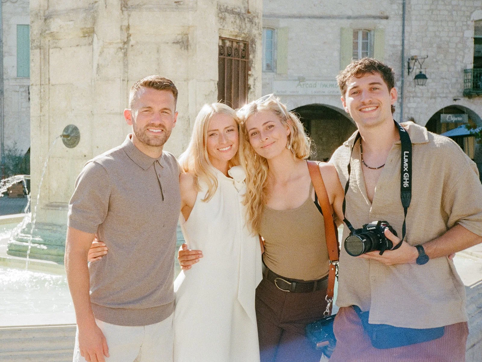 Group of four young adults smiling outdoors near a fountain in EyMet, South Of France, in a European town. One couple is the bride and groom and the other is the filmmaking team.