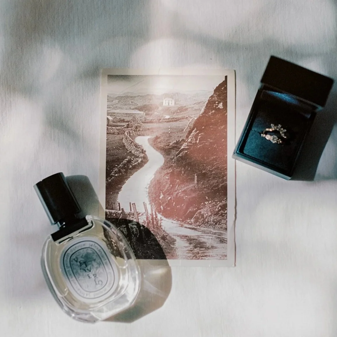 A black and white landscape photo of a winding river in a valley with hills and rocks, placed on a white surface. To the left, there is a bottle of perfume, and to the right, a jewelry box with rings inside. Image is a wedding flatlay of details.