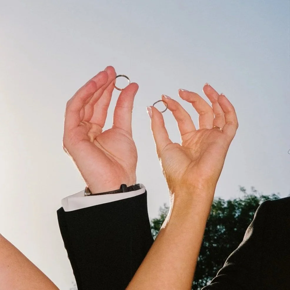 Two hands holding wedding rings outdoors against a bright sky and trees.