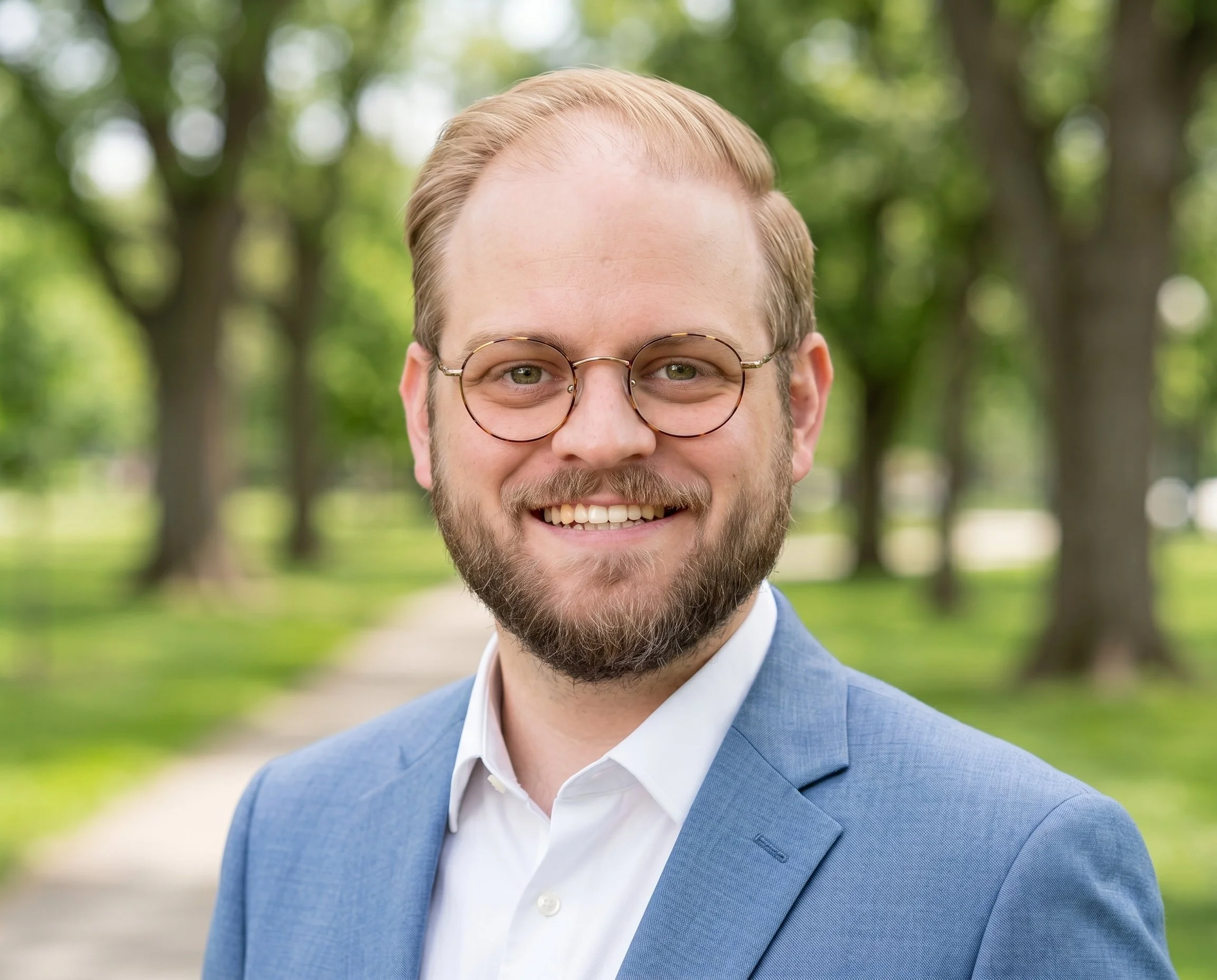 A smiling man with blond hair, beard, and glasses, wearing a light blue suit jacket and white shirt, standing outdoors in a park with green trees in the background.
