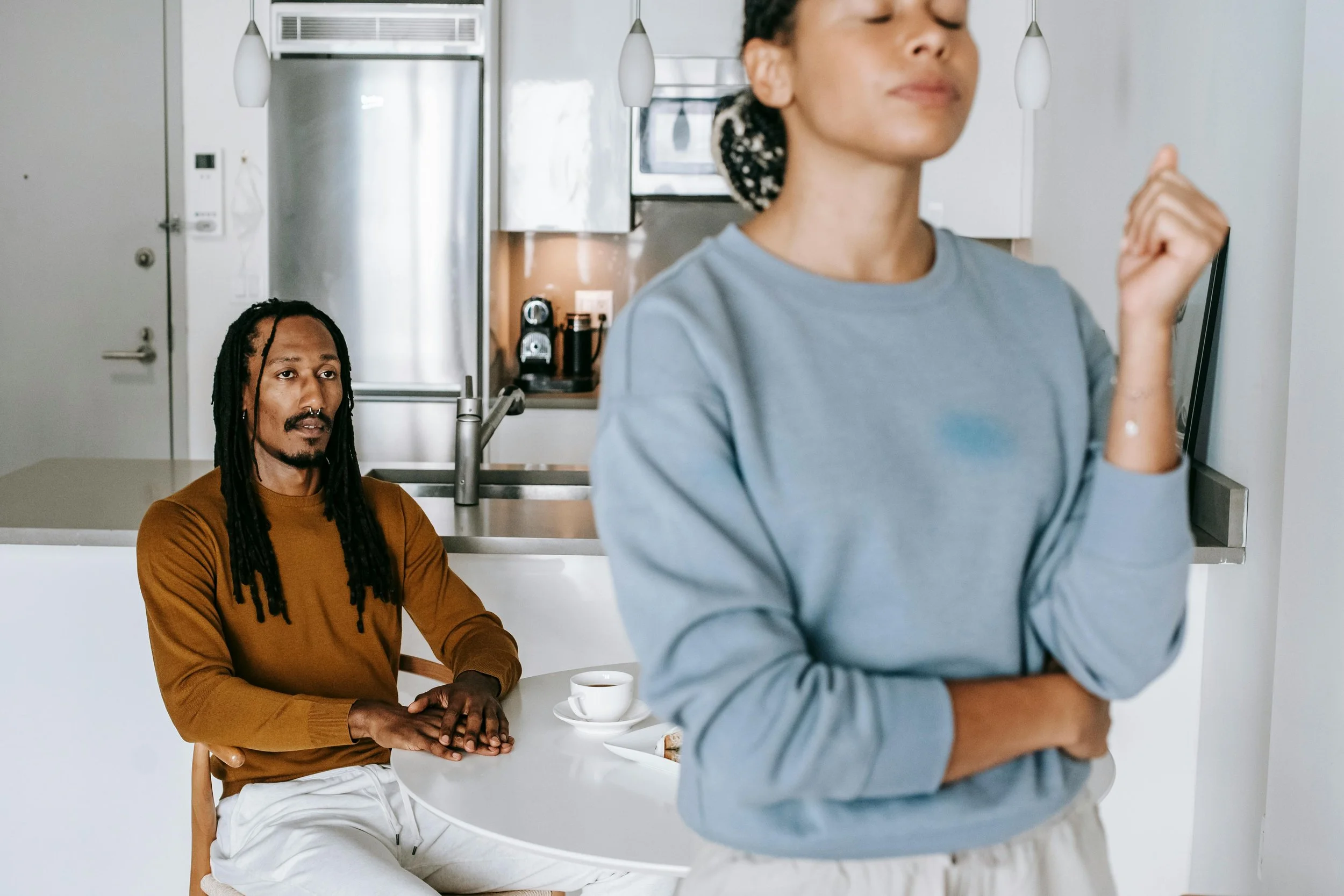 A woman with dark, curly hair in a bun is in the foreground with eyes closed and hand in a fist. A man with long dreadlocks, a beard, and mustache, wearing a brown shirt, is seated at a white kitchen counter in the background, looking at the woman, with coffee cups and a plate with a pastry on the counter.
