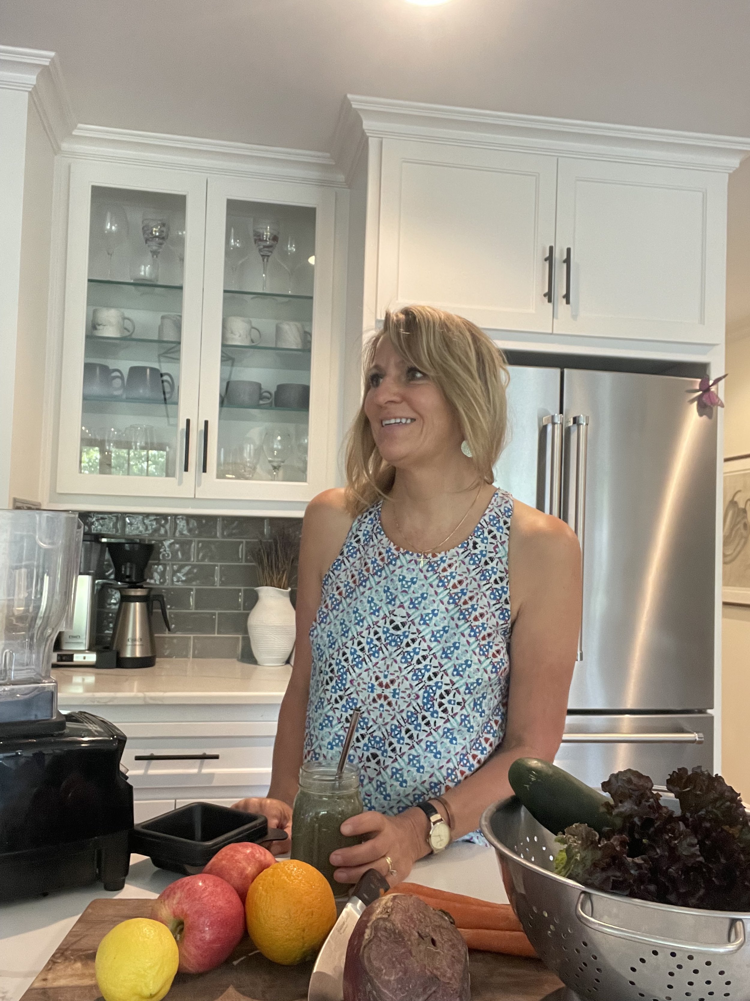 A woman standing in a modern kitchen, smiling and holding a jar of green smoothie. The kitchen features white cabinets, a stainless steel refrigerator, and various fresh fruits and vegetables on the counter.