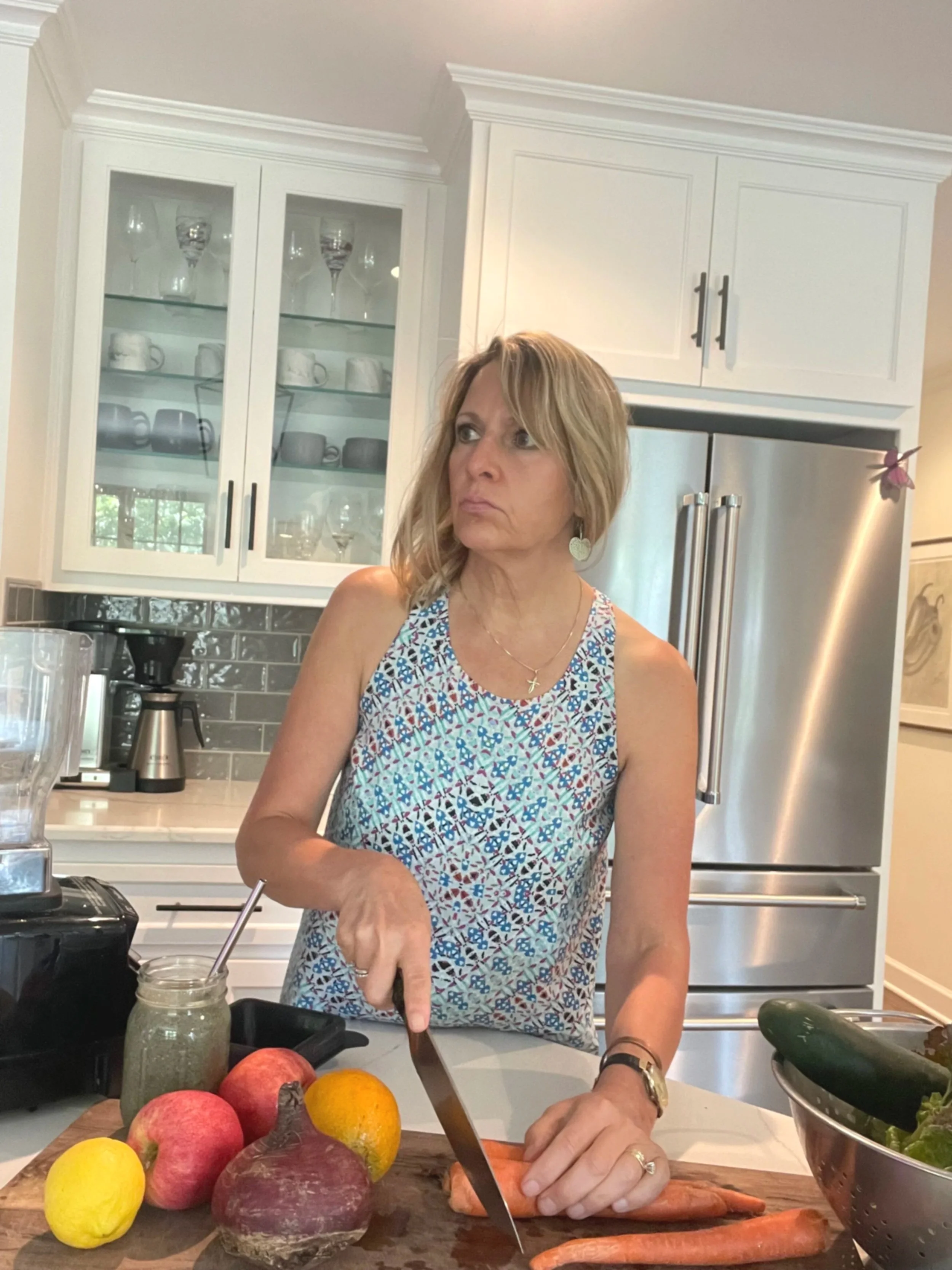 A woman in a patterned sleeveless top cuts carrots in a kitchen with white cabinets, a stainless steel refrigerator, and a grey tiled backsplash. There are apples, a lemon, an orange, and a jar of green smoothie on the counter, with a bowl of zucchinis nearby.