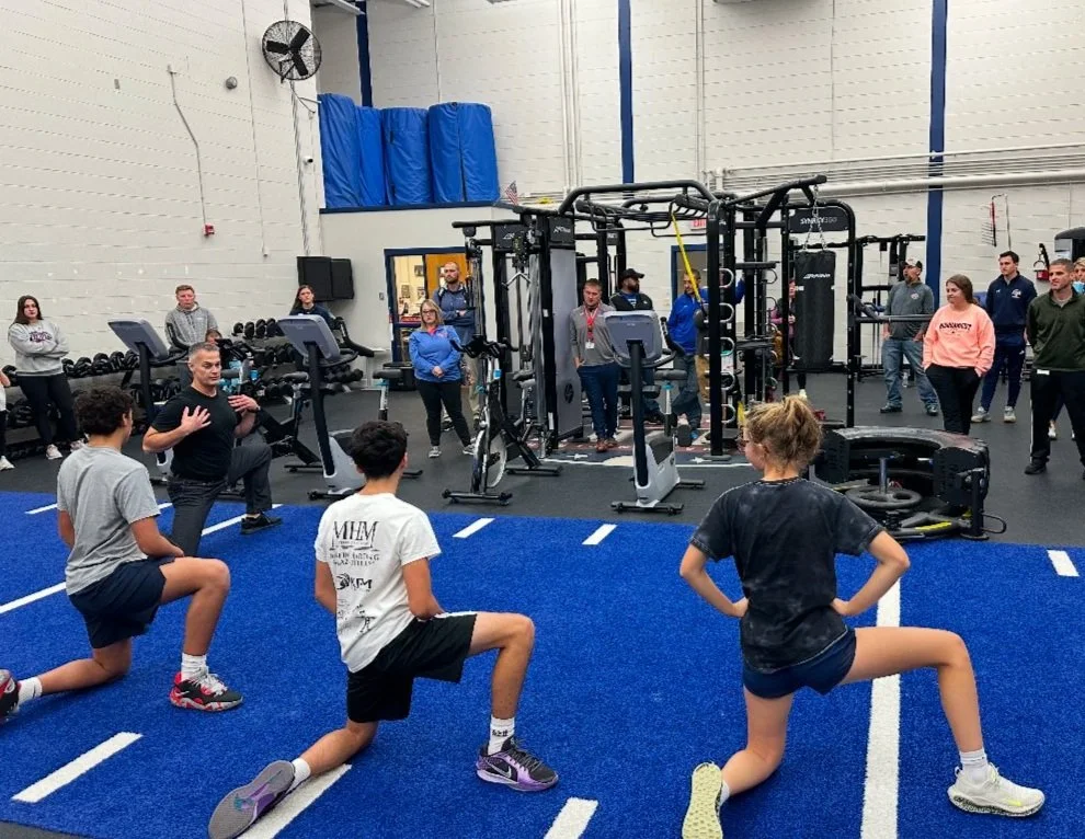 A fitness coach demonstrating exercises to four kids doing lunges on a blue mat in a gym, with adults watching in the background.