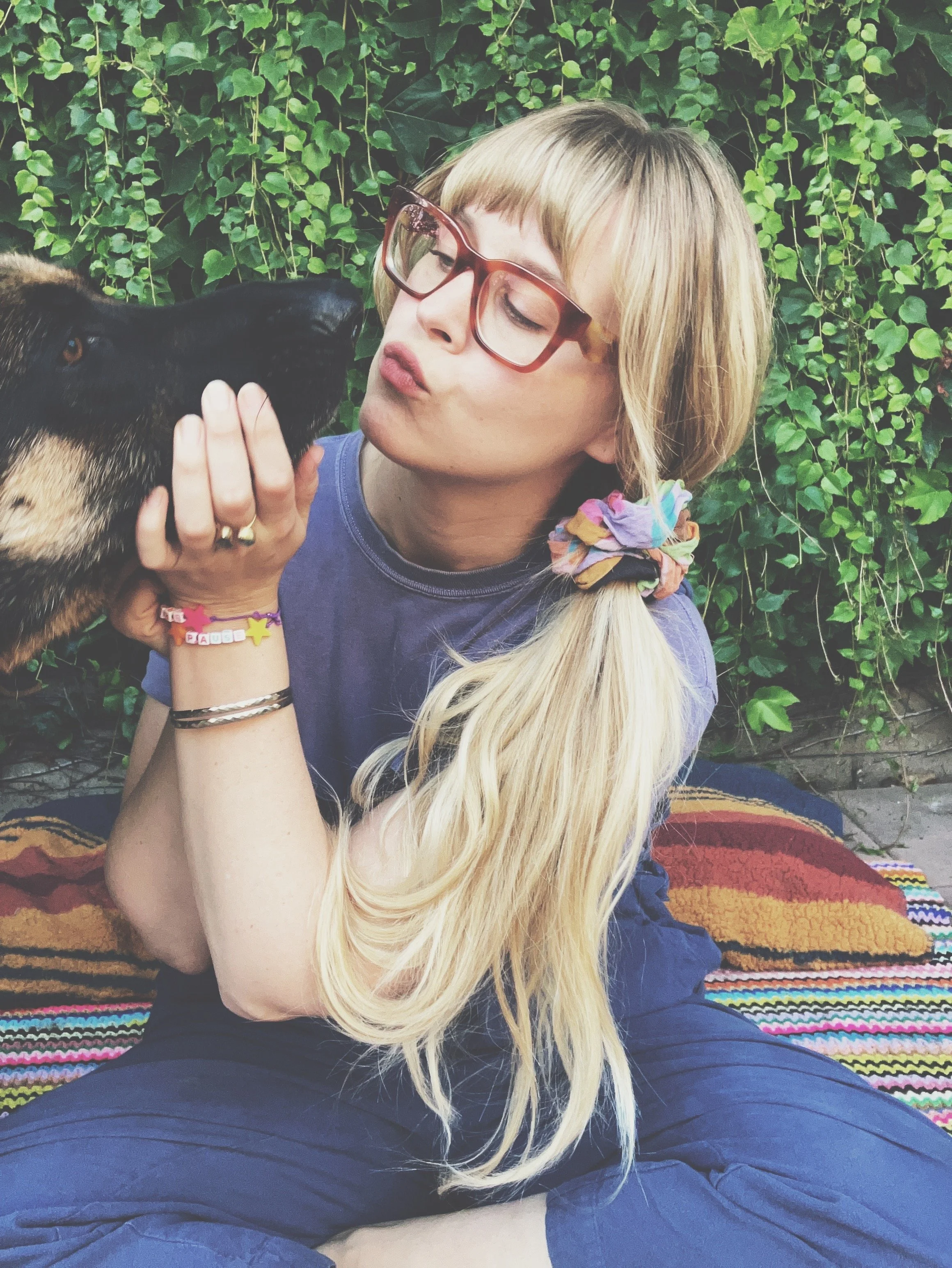 A woman with long blonde hair, wearing glasses and colorful bracelets, is sitting on a multicolored blanket outdoors, holding a black dog close and giving it a kiss.