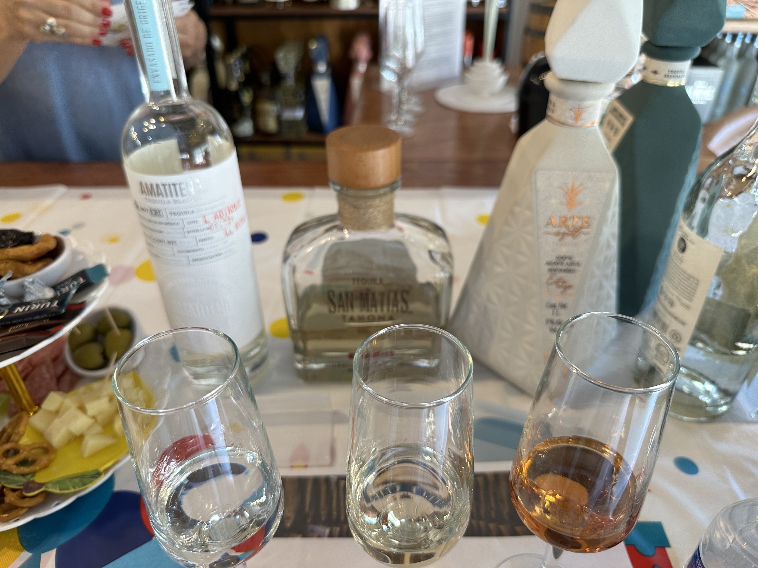 A table with various bottles of liquor, three empty glasses, and a glass with rosé wine. There are snacks and a person in the background.