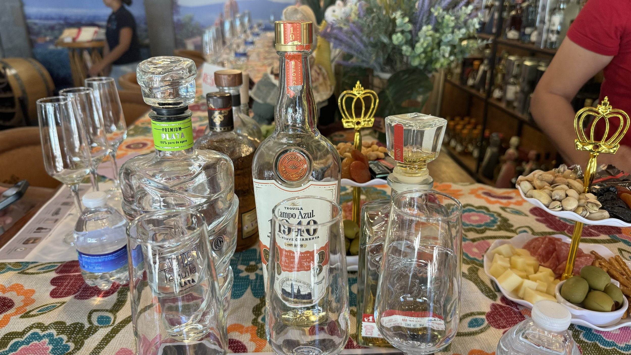 Table with liquor bottles, wine glasses, water bottles, and dishes with cheese, olives, nuts, and cured meats, set on a colorful embroidered tablecloth in a restaurant or bar setting.