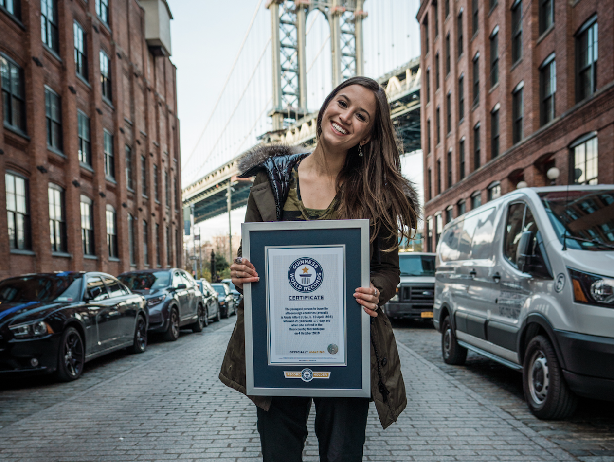 Young woman standing on city street holding a Guinness World Records certificate, with cars and bridges in the background.