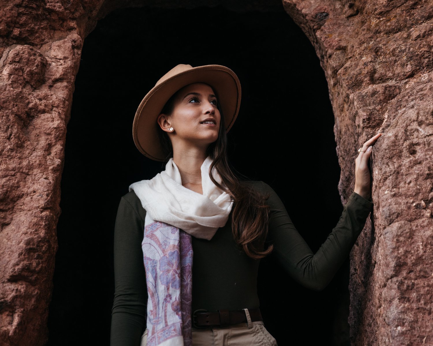 Lexie Limitless, with a wide-brimmed hat and a scarf around her neck, standing in a rocky cave opening, looking up and to the side, with her hand touching the rock wall.