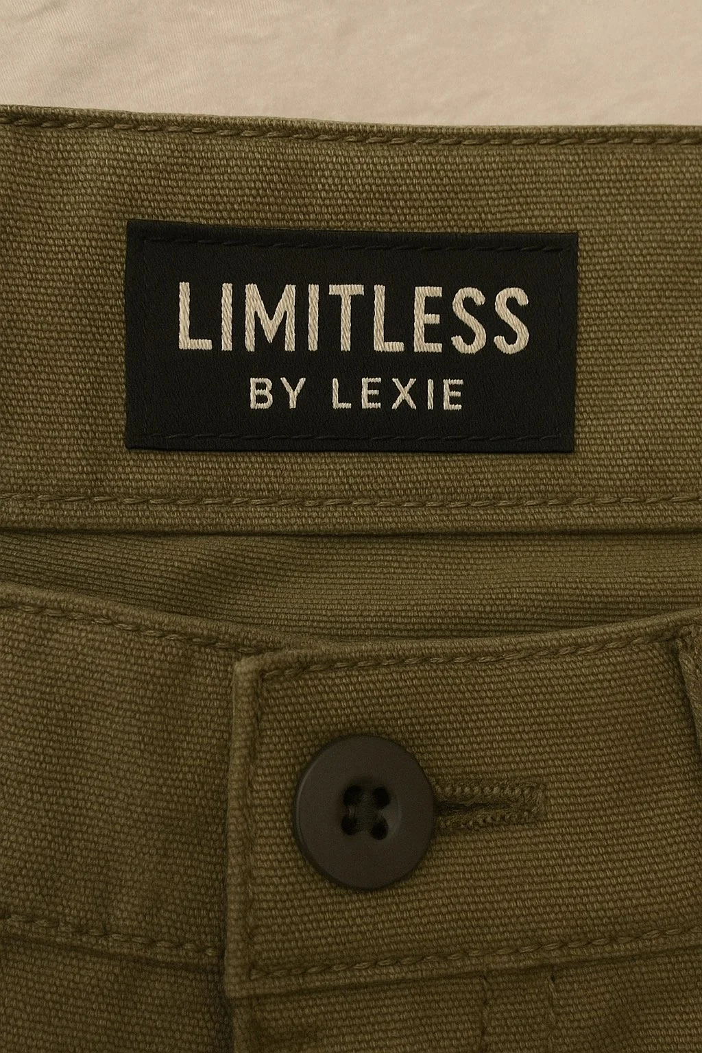 Close-up of a khaki-colored fabric with a black label that reads 'LIMITLESS BY LEXIE' sewn onto it, and a black button with four holes visible below the label.
