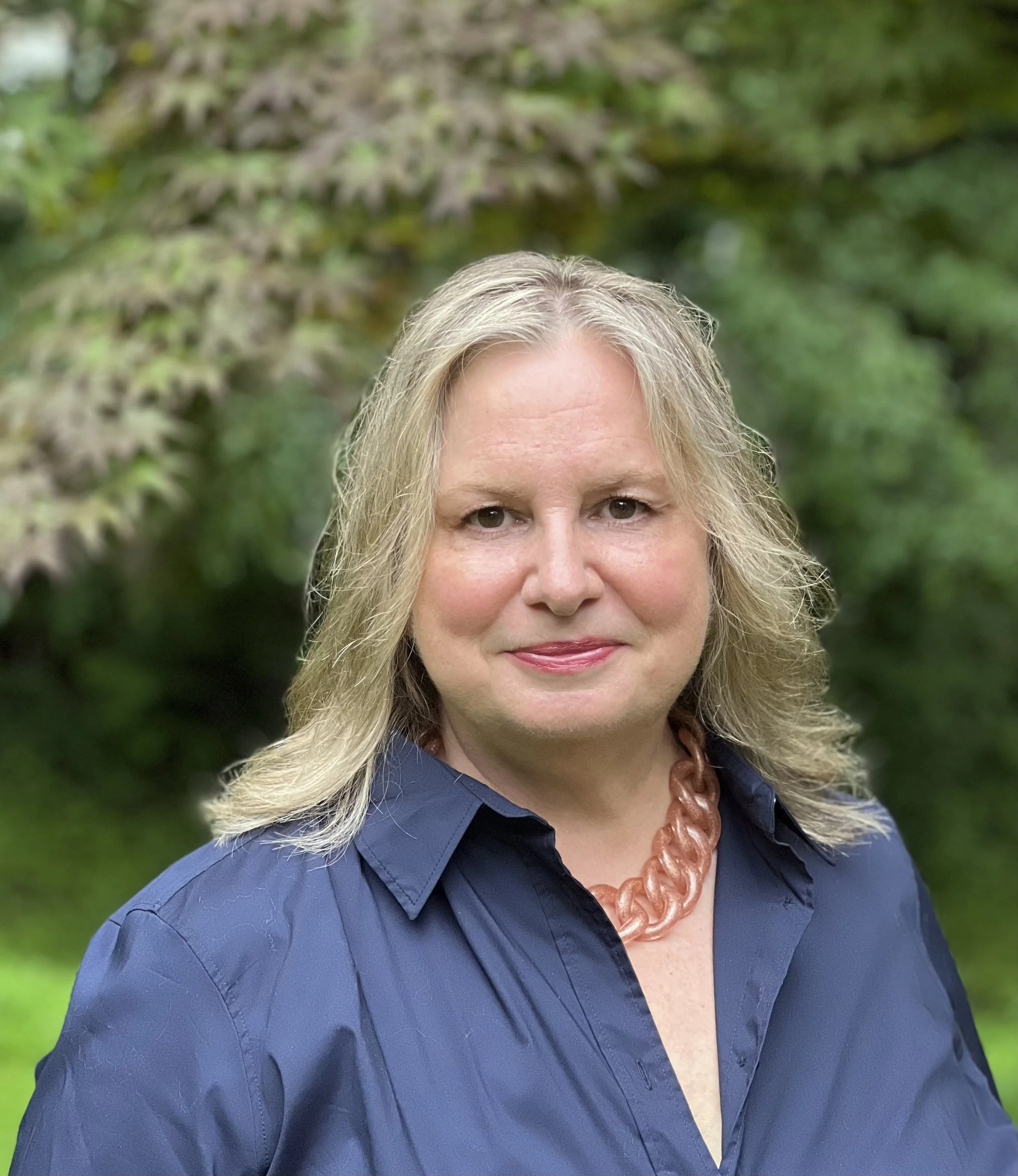 Close-up of a smiling middle-aged woman with blonde hair, wearing a navy blue shirt and a chunky pink necklace, standing outdoors with green trees in the background.