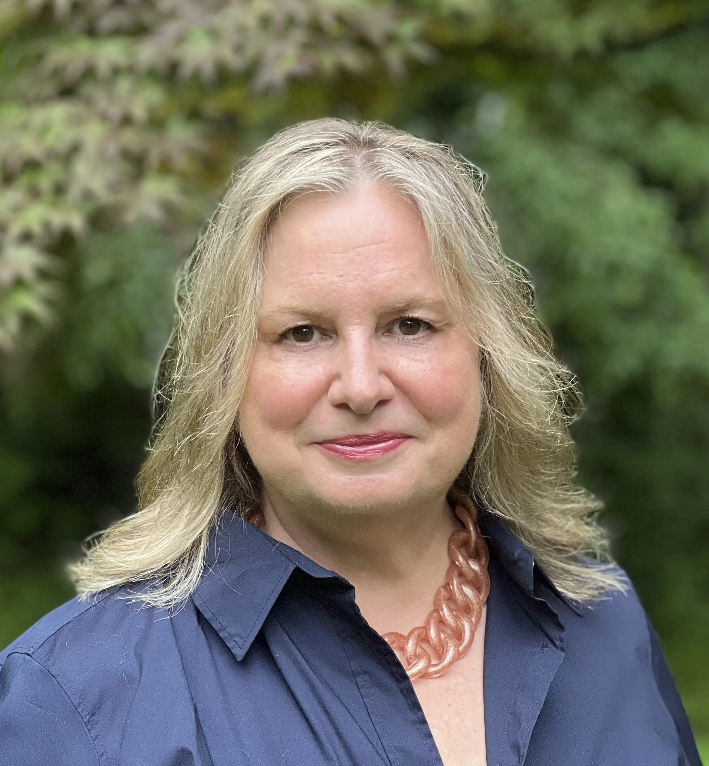 Close-up of a woman with blonde hair, wearing a blue shirt, a brown jewelry necklace, and standing outdoors with blurred green foliage in the background.