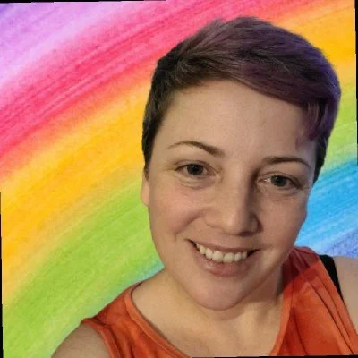 Person with short dark hair smiling in front of a rainbow painted wall, wearing an orange sleeveless top.
