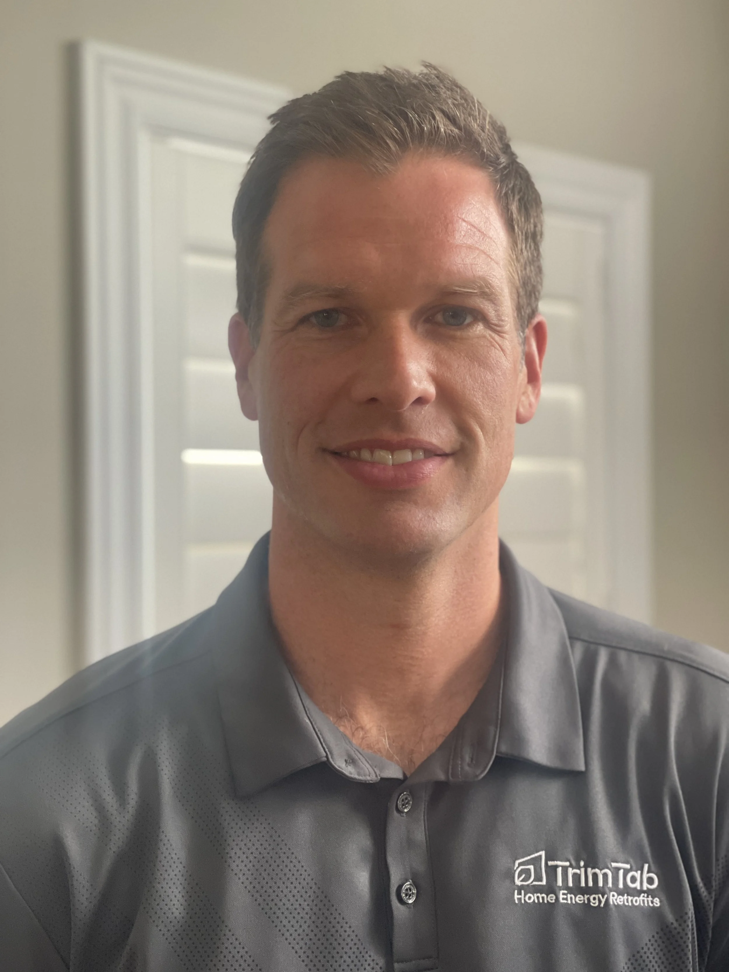 A man with short brown hair, wearing a gray polo shirt with a company logo, standing indoors in front of a door with white blinds.