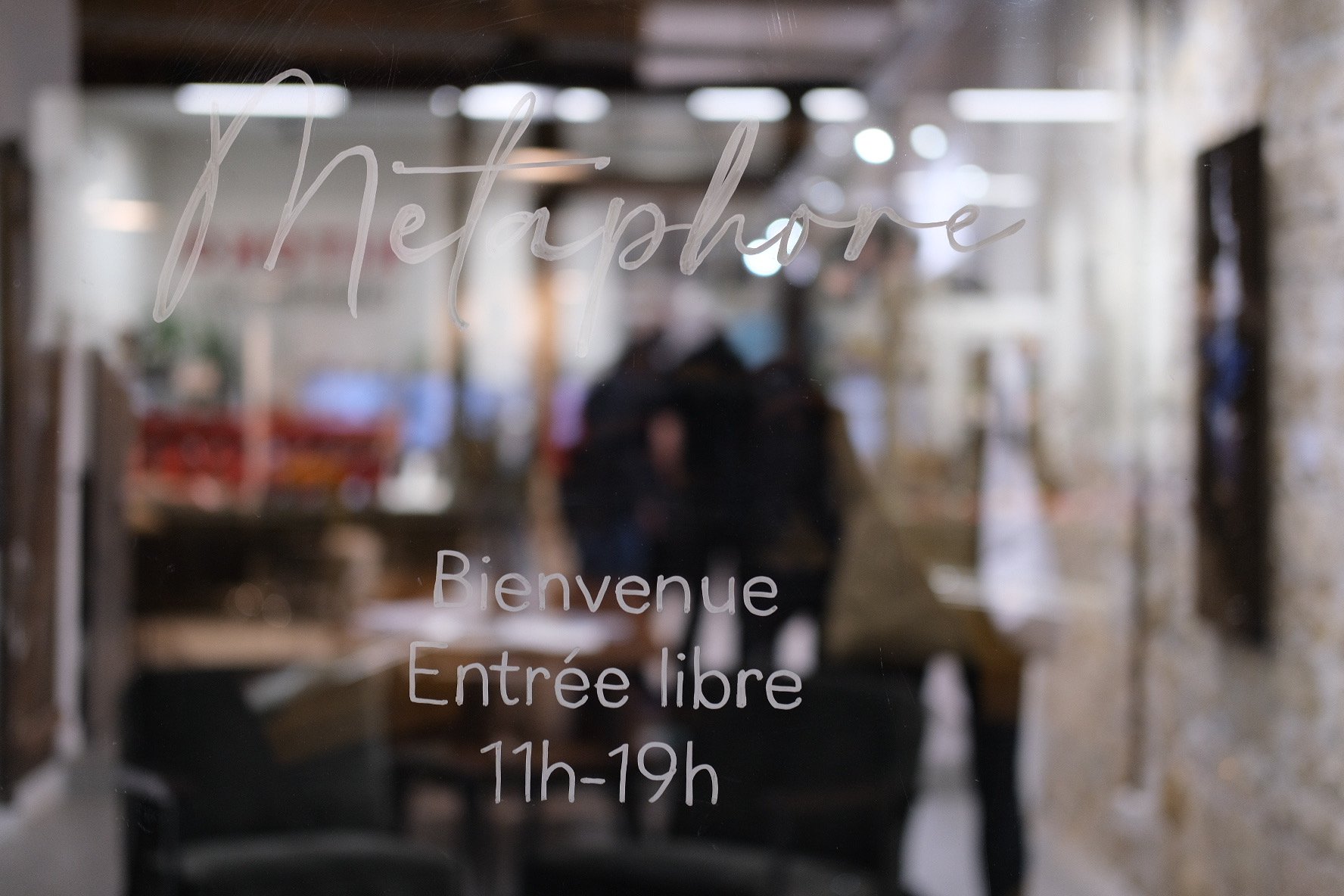 View through a glass door with handwritten sign displaying the restaurant's name, opening hours from 11 a.m. to 7 p.m., and welcoming message in French, with blurred interior and people inside visible behind the glass.