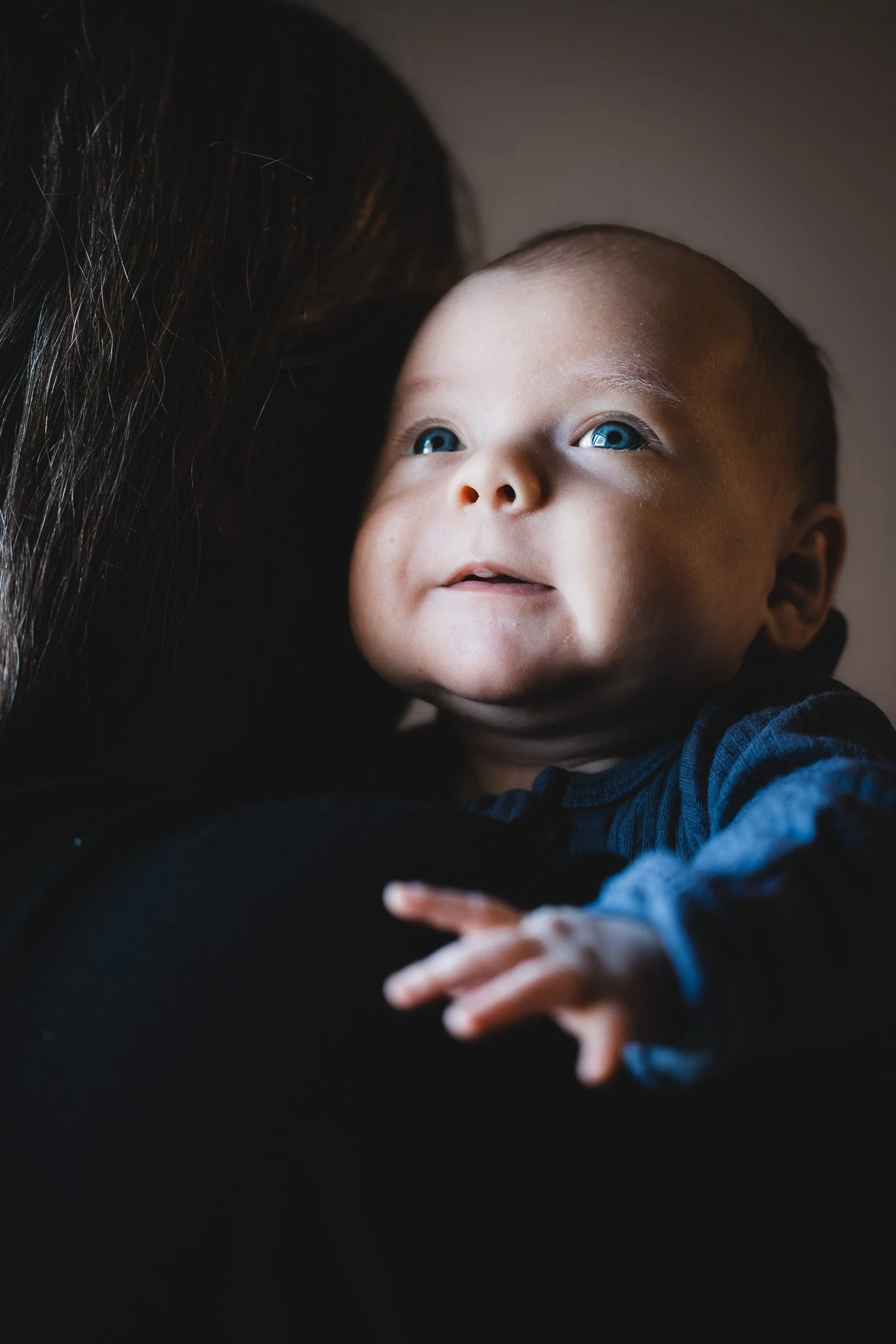A baby with blue eyes looking up while being held against a dark background.