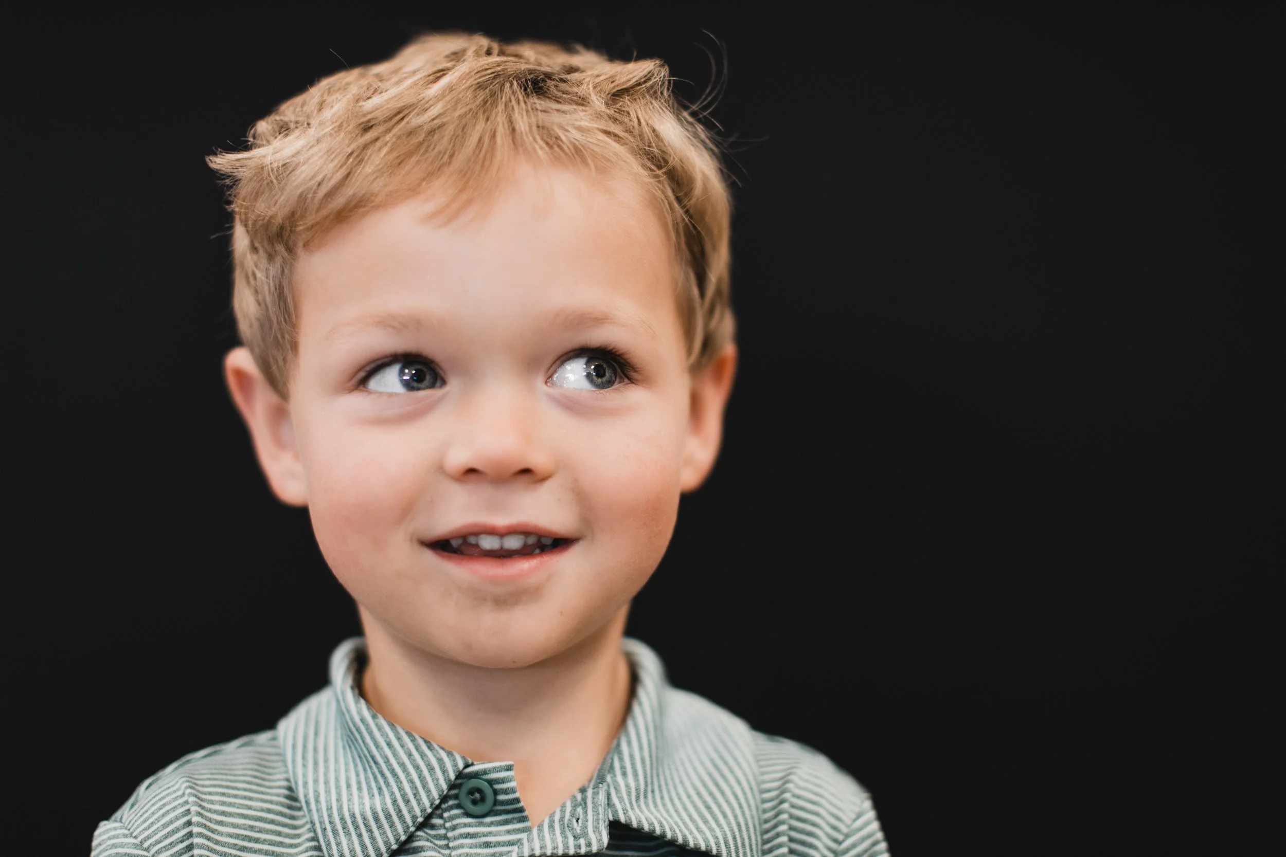 A young boy with blonde hair and blue eyes wearing a striped collared shirt, smiling and looking to his right against a black background.