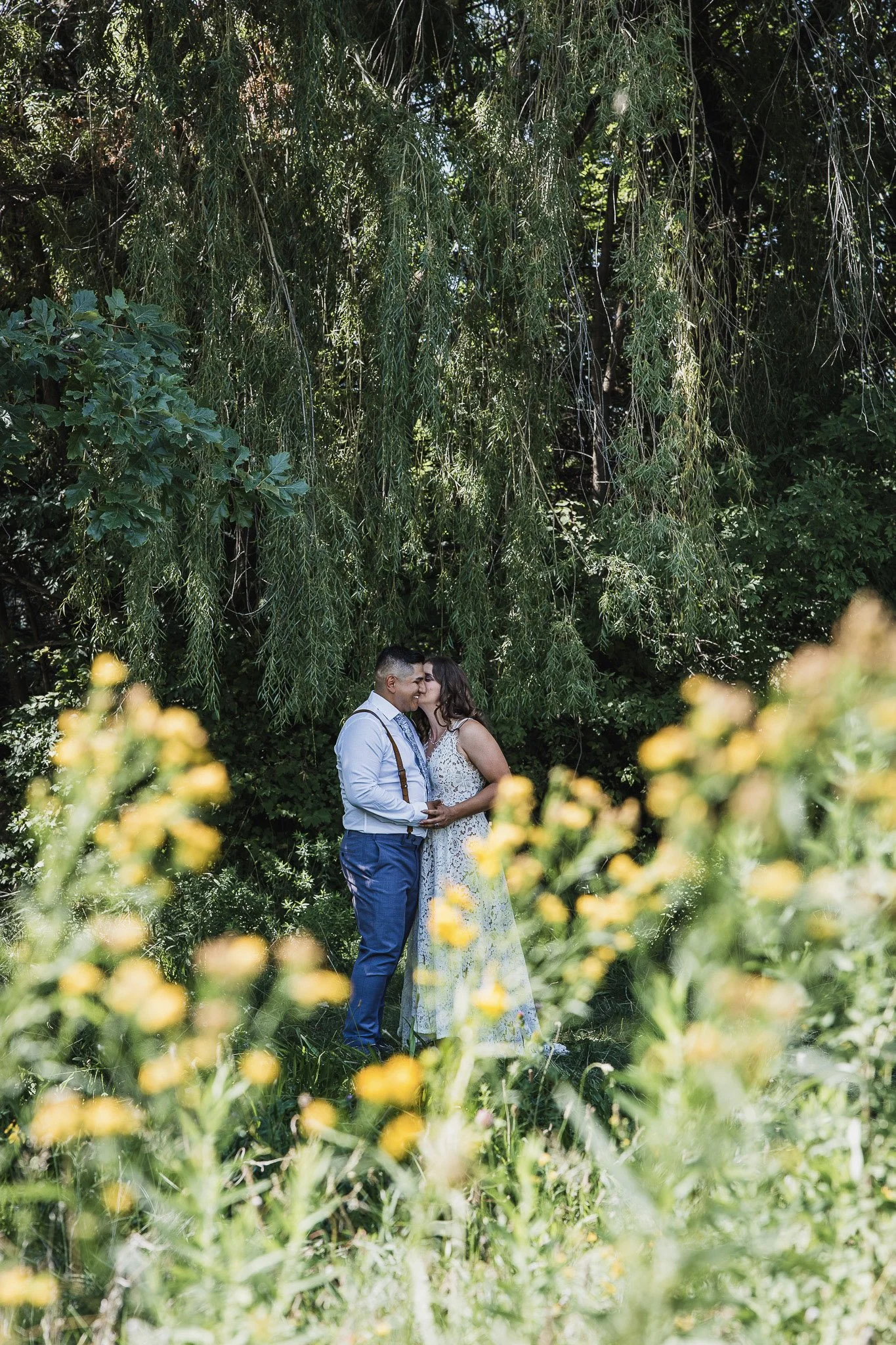 A couple sharing a kiss in a lush green garden, surrounded by trees and yellow flowers.